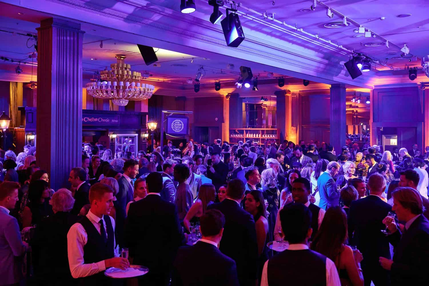 A large group of people in formal attire socialize at the London Press Club's bustling indoor event, the Press Club Ball, with ambient purple and orange lighting.