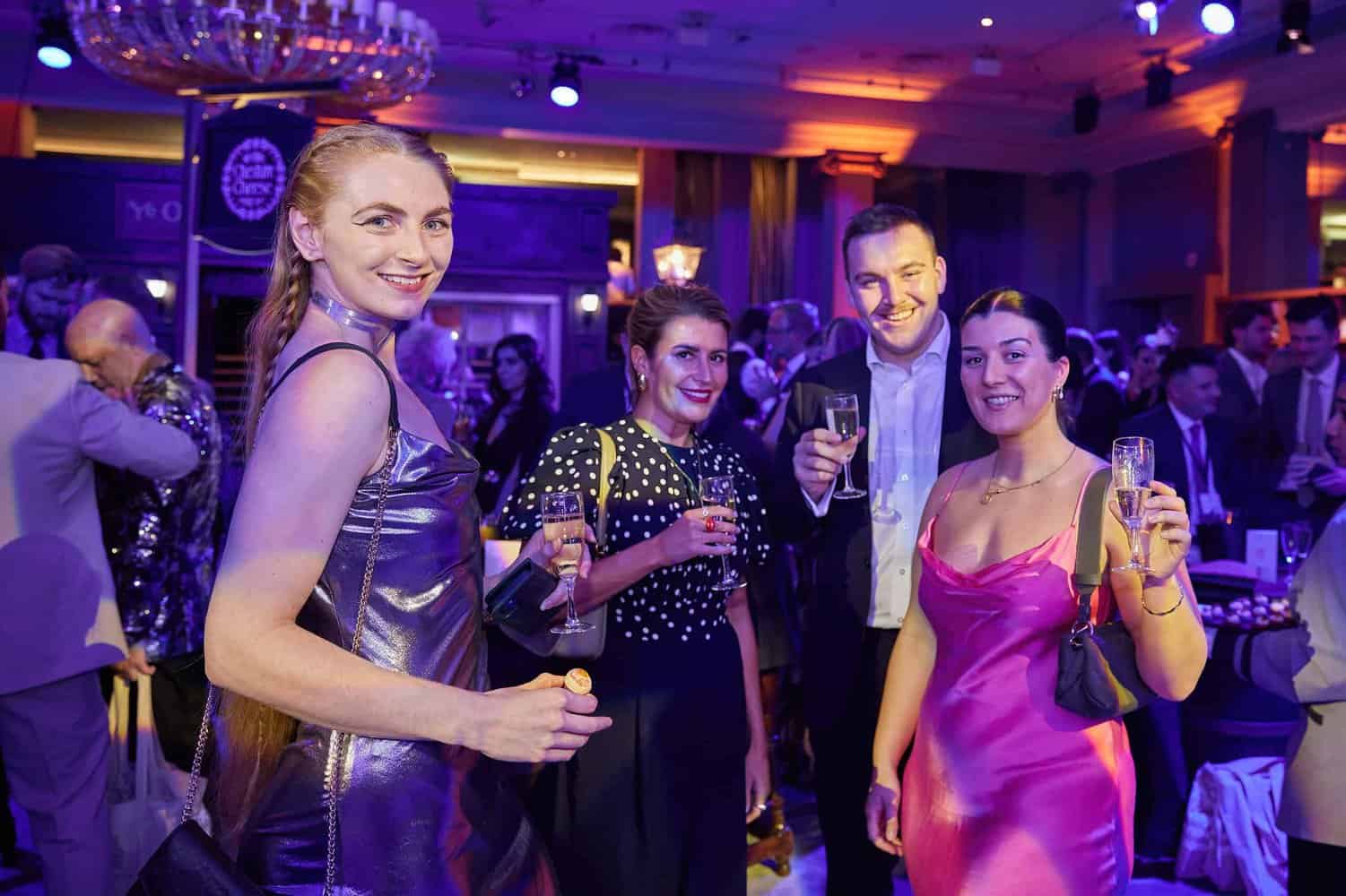 Four people in formal attire hold champagne glasses at a lively indoor event with colorful lighting, reminiscent of the vibrant atmosphere at the London Press Club's annual gala.