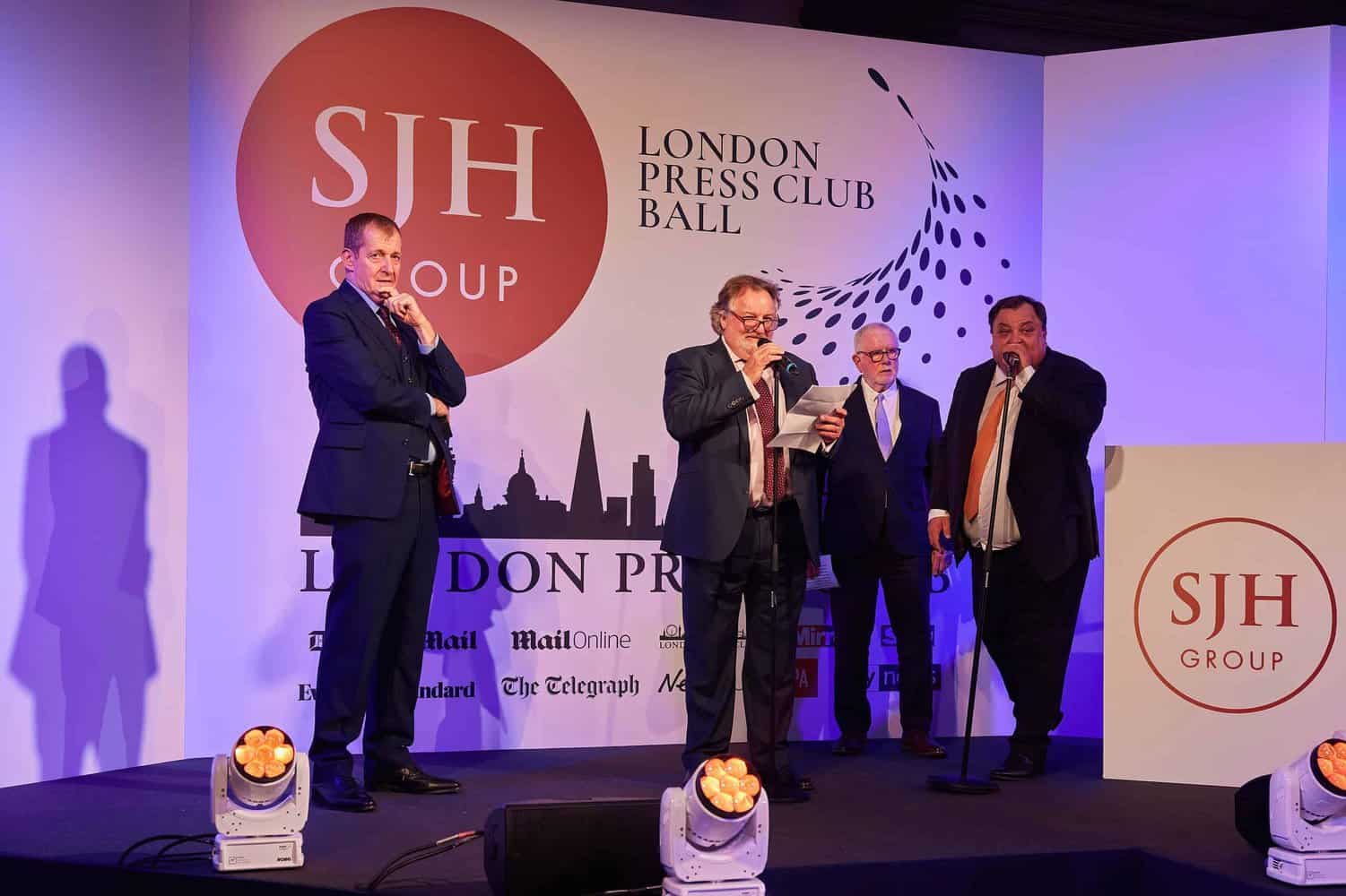 Four men in suits stand on a stage with microphones at the London Press Club Ball, a prestigious London event, in front of a backdrop featuring logos of various media outlets.