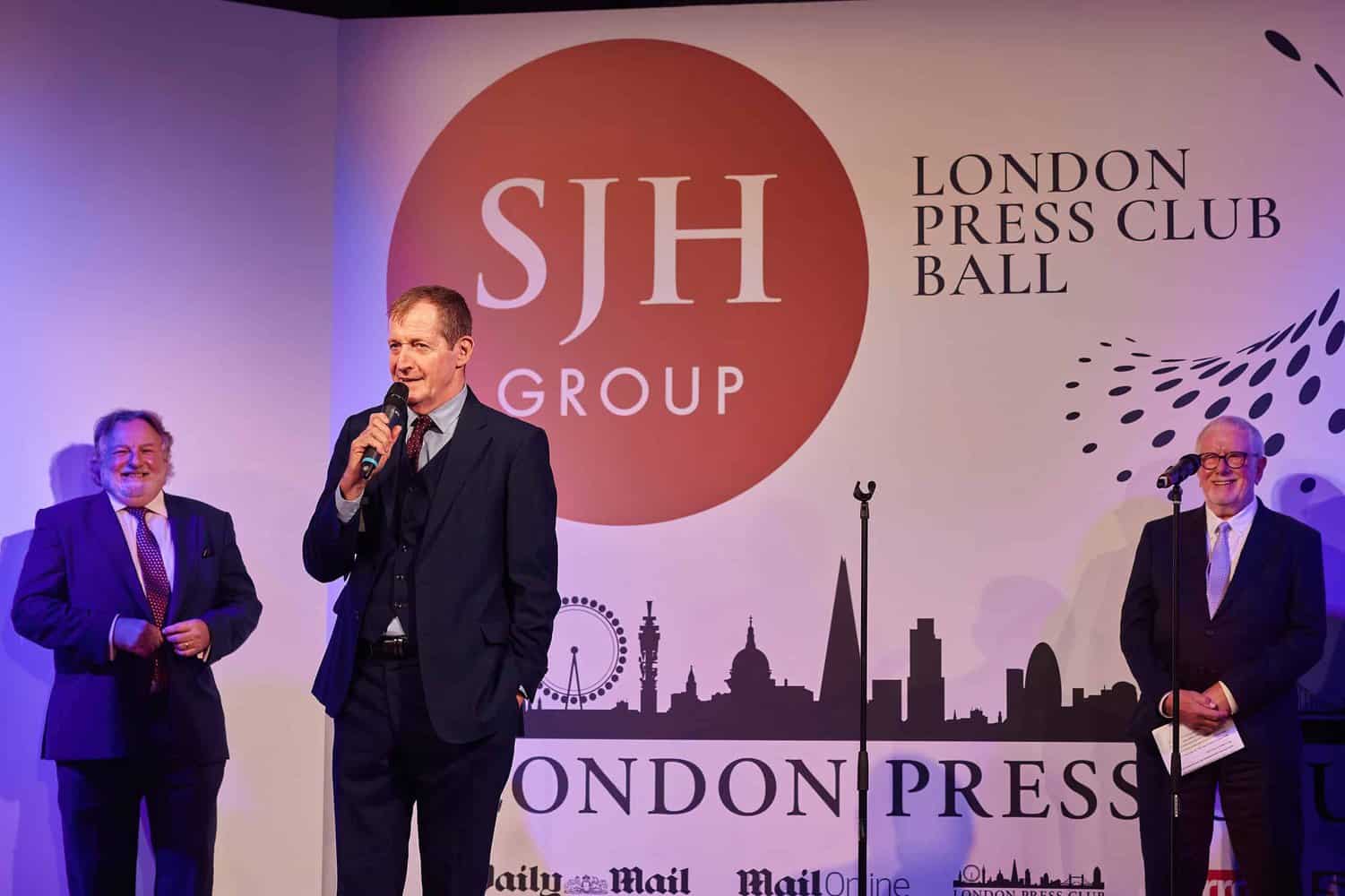 Three men in suits take center stage at the prestigious London Press Club Ball, with one eloquently speaking into a microphone.