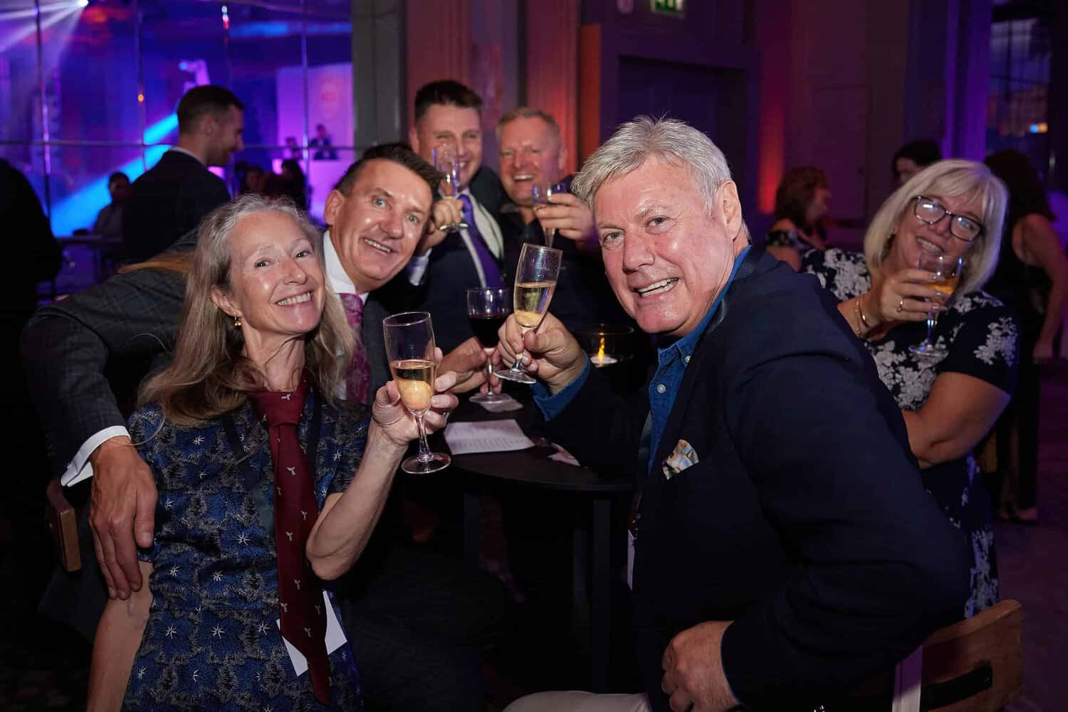 In a dimly lit room, a group of people at the Press Club in London sit around a table, smiling and holding glasses filled with drinks, enjoying the jovial atmosphere akin to a lively ball.