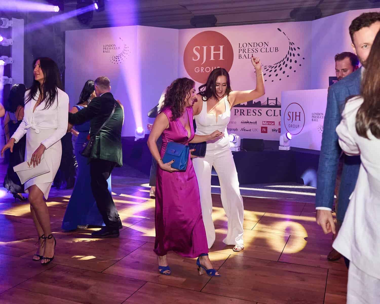 People dancing at a lively London Press Club Ball, with vibrant signage in the background. Women in colorful dresses and men in suits twirl on the dance floor, embodying the electrifying spirit of the event.