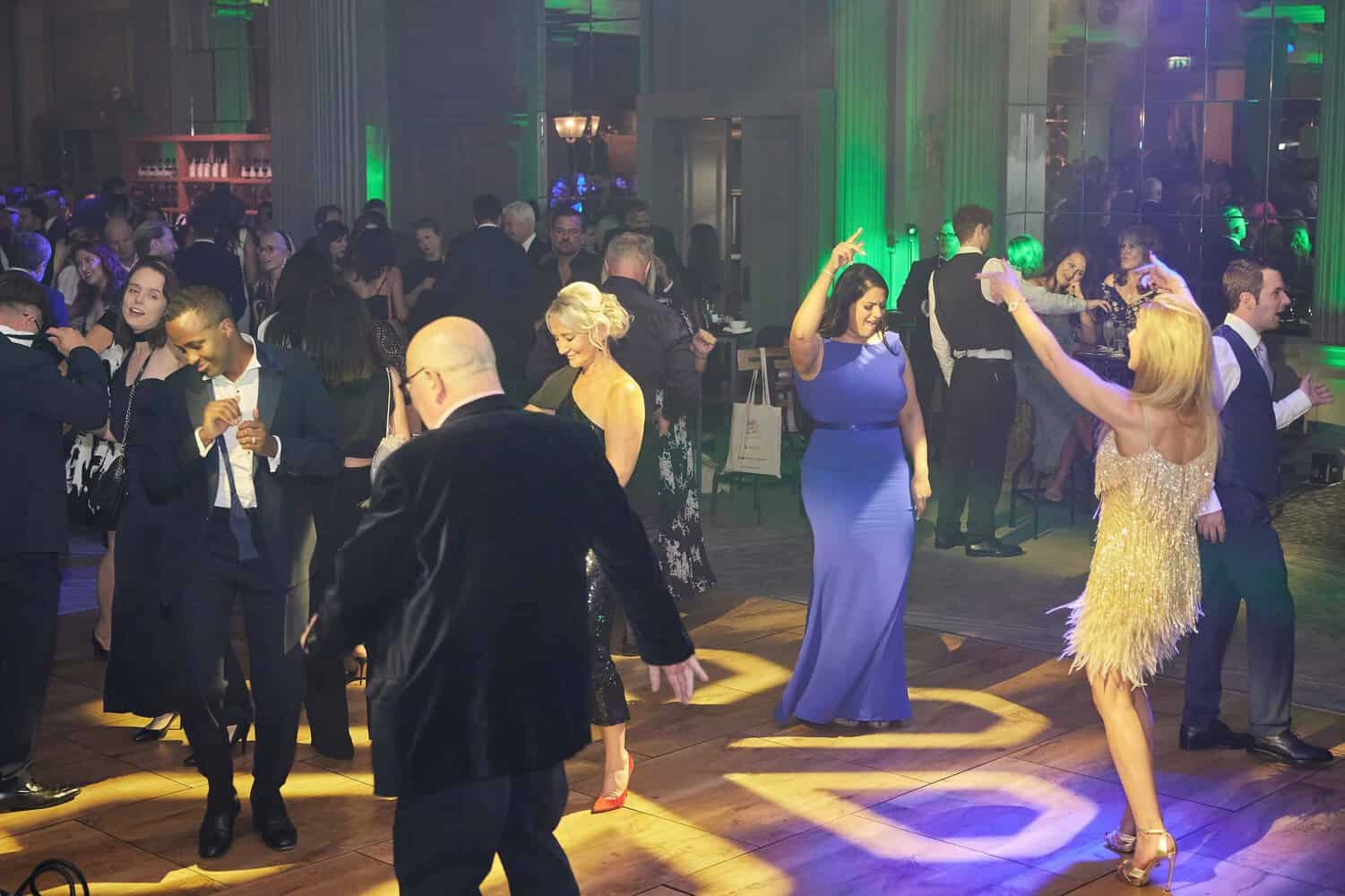 Amidst the vibrant green lighting of the London Press Club, people in formal attire dance energetically on a wooden floor, bringing life to the elegant ball.