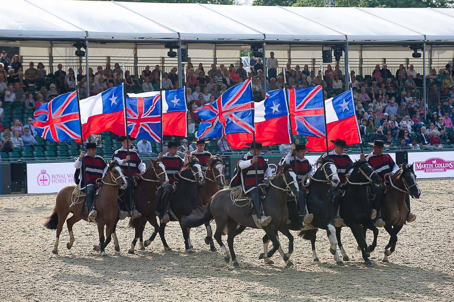 The equestrian team performs with elegance, holding blue and red flags before an audience under a covered stand, reminiscent of a scene from a royal documentary celebrating the Queen's 90th birthday.