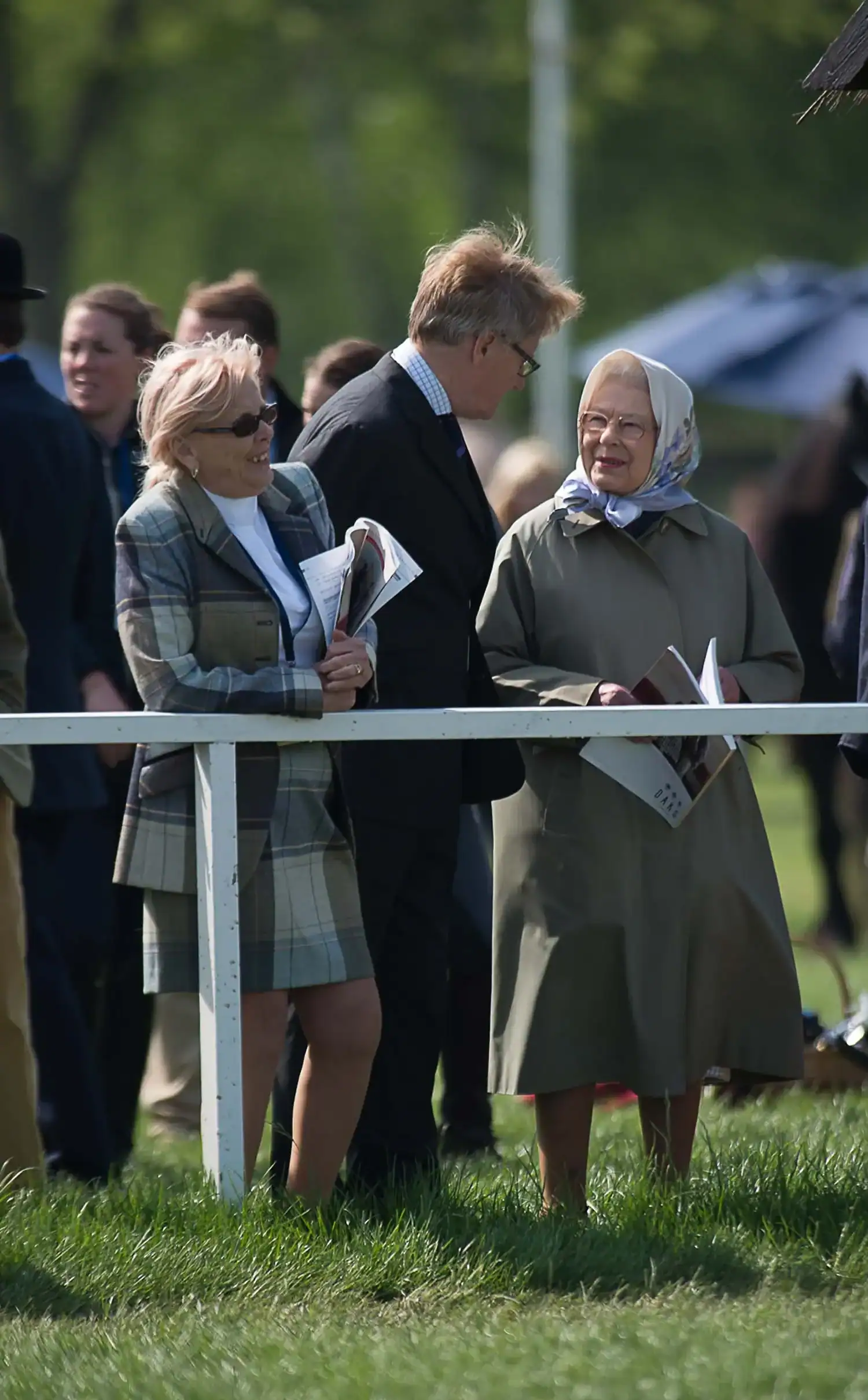 Three people stand outdoors at a railing, dressed in formal attire, holding papers. A woman wearing a headscarf smiles at a man in a suit and a woman in a plaid coat, reminiscent of the elegance seen during the Royal Family's celebrations for the Queen's 90th Birthday.