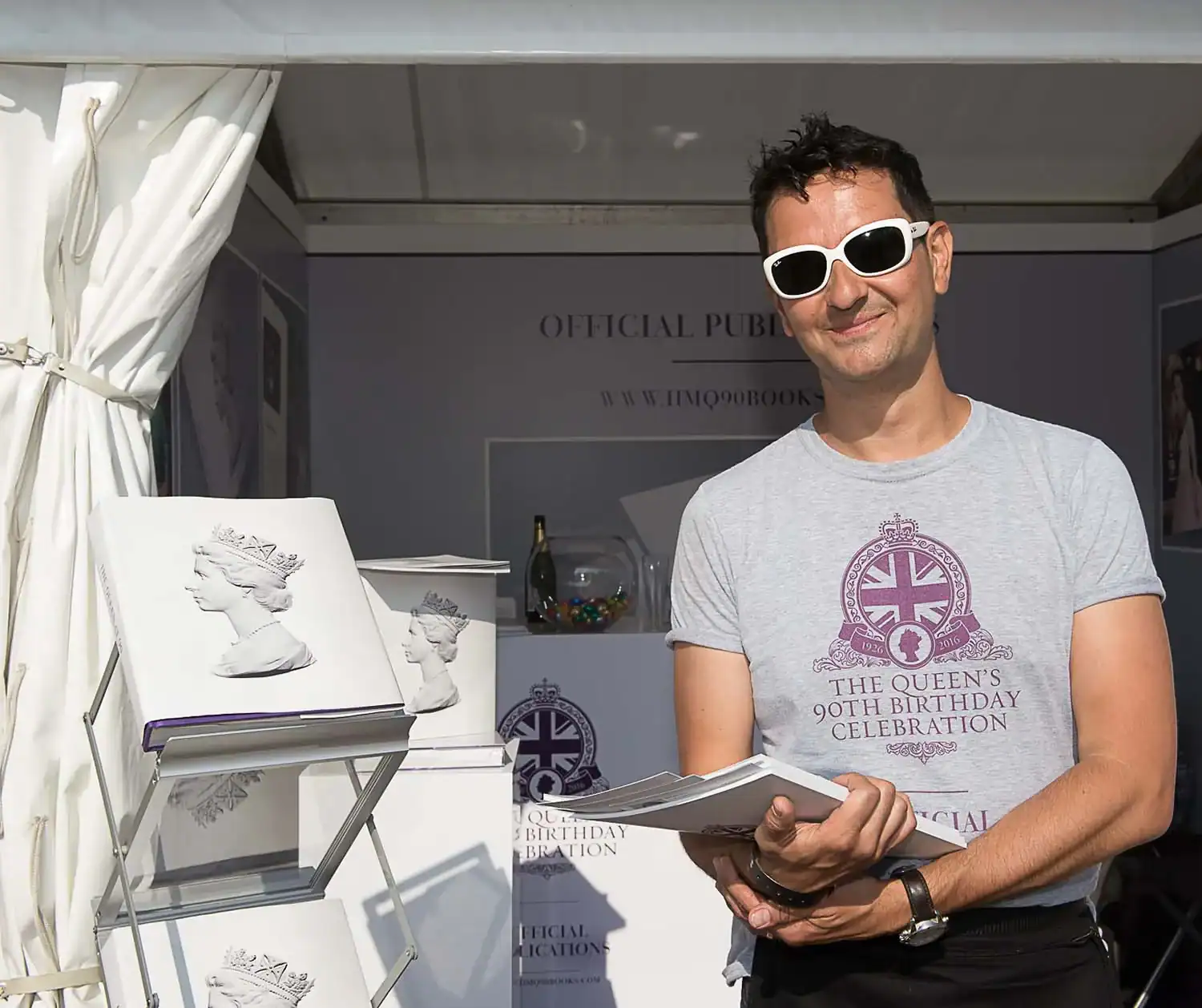 A person wearing sunglasses and a "The Queen's 90th Birthday Celebration" shirt stands beside a display of commemorative books in a booth, capturing the essence of the royal celebration.