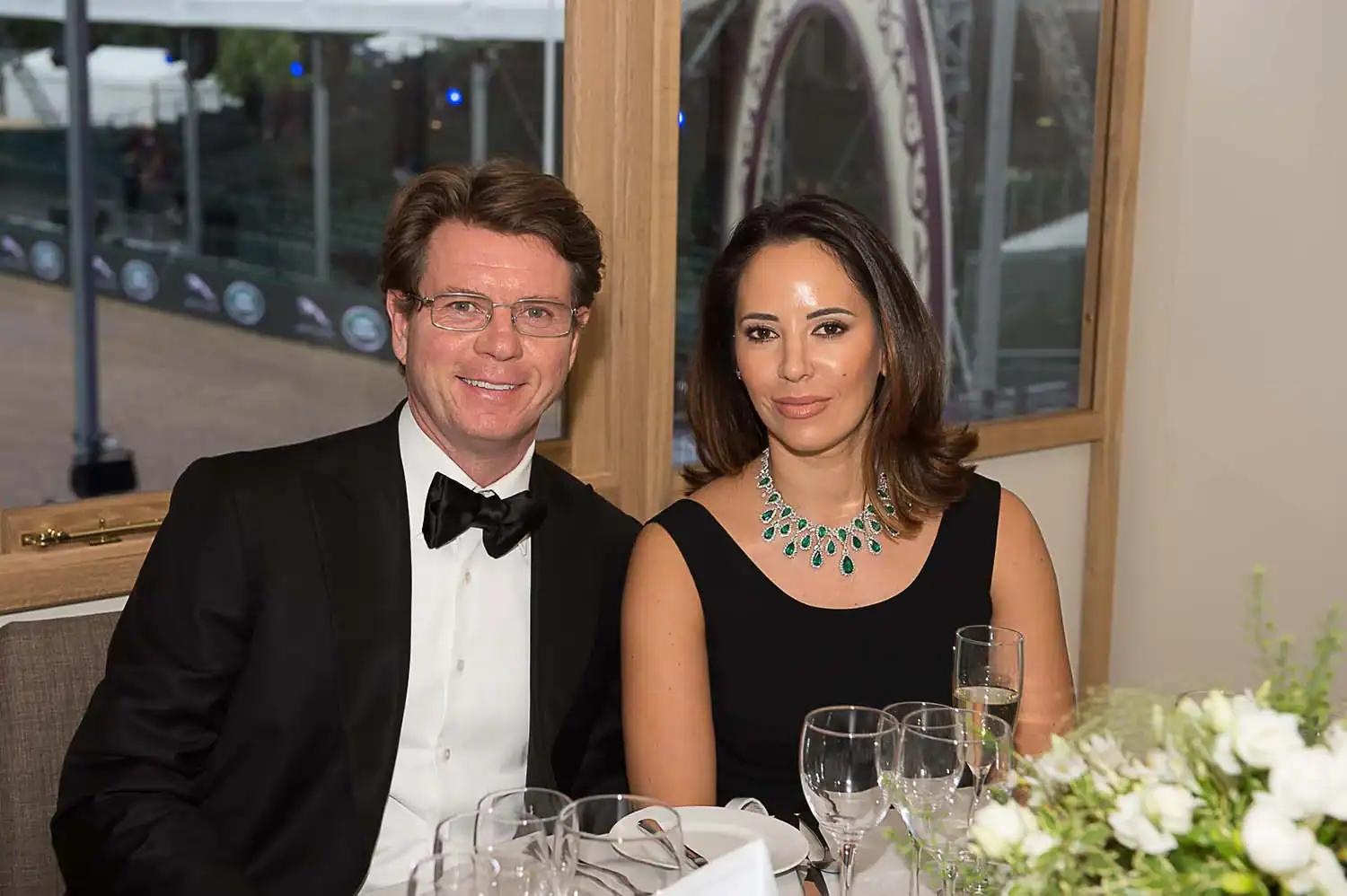 A man in a black tuxedo and a woman in a black dress adorned with a green necklace sit together at a table set for a formal event, reminiscent of the elegance celebrated during Queen Elizabeth II's 90th birthday.