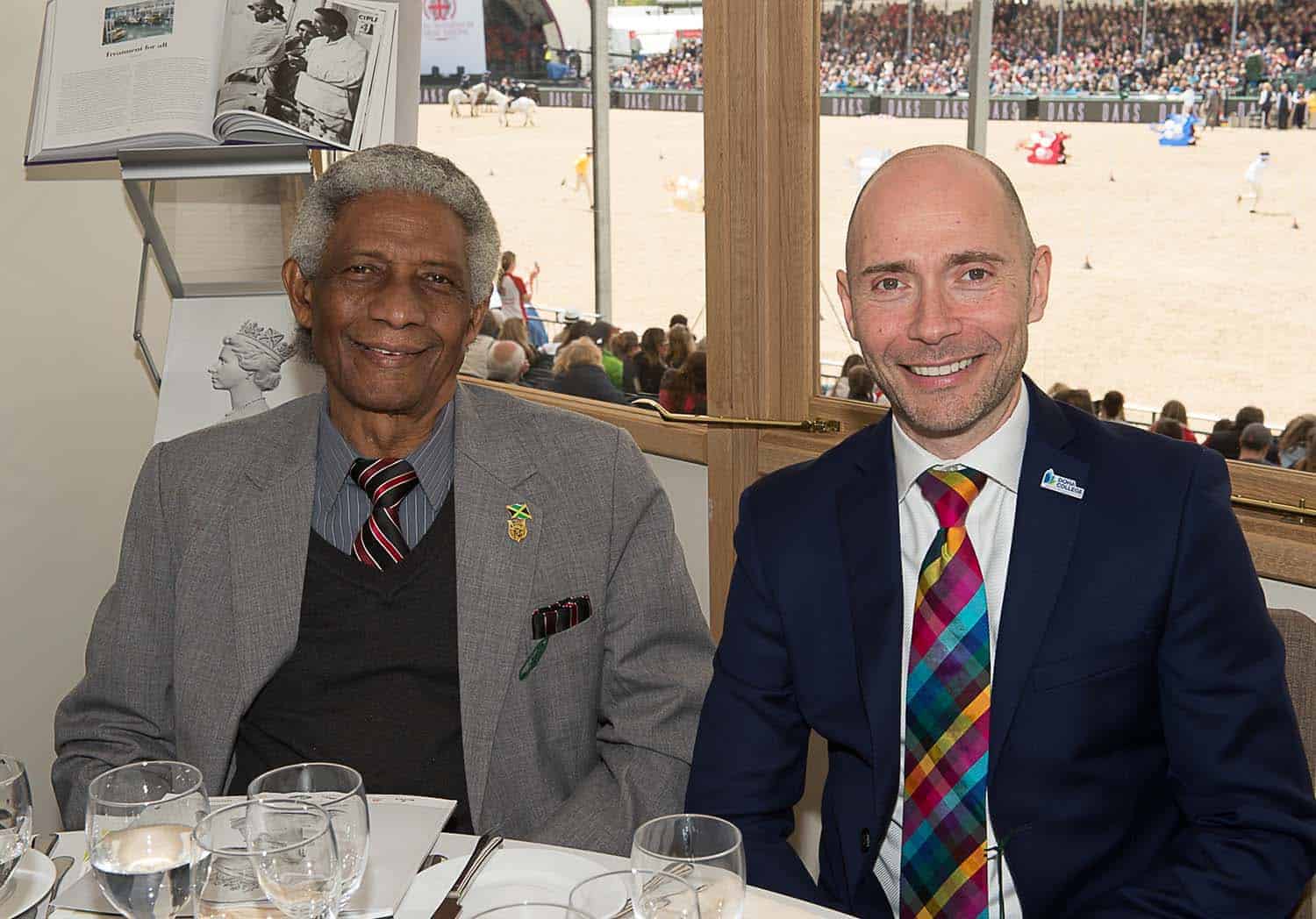 Two men in suits sit at a table with wine glasses, smiling, as a royal celebration unfolds. A horse event is visible outside through the window, adding to the festive atmosphere befitting a 90th birthday tribute.