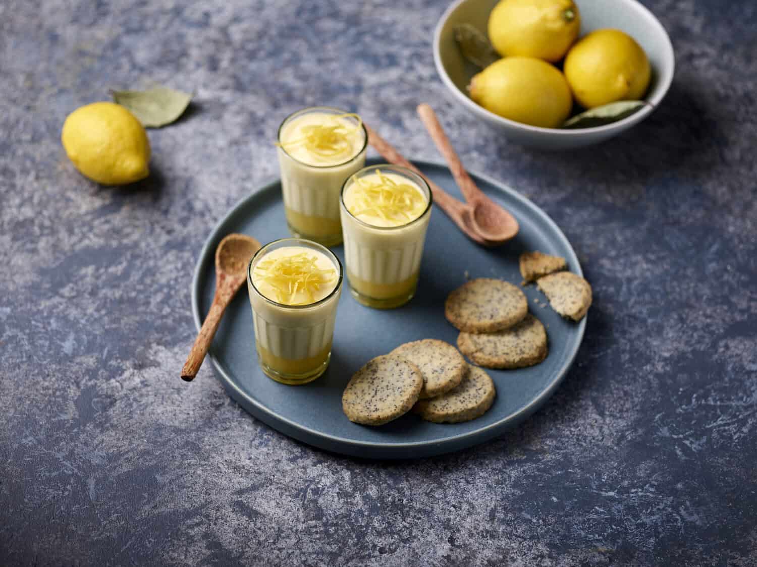 Three glasses of lemon dessert with spoons and cookies on a plate. Lemons in a bowl and one lemon outside on a textured surface, waiting to be transformed by the heroic art of cooking.