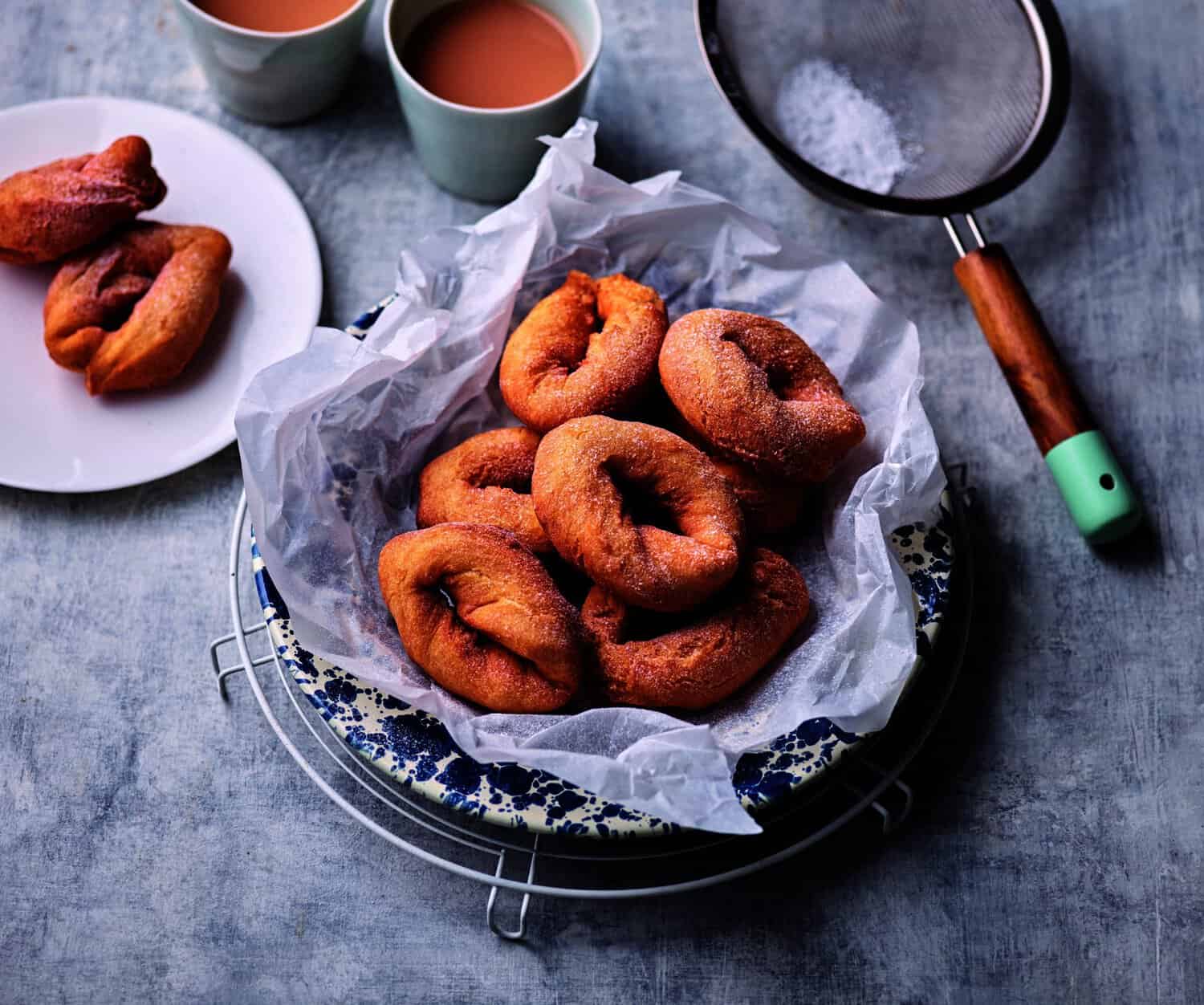 A plate with fried pastries on parchment paper, a sieve, and two cups of tea on the side—truly a cooking hero's delight. A separate white plate holds additional pastries, ready to save any afternoon craving.