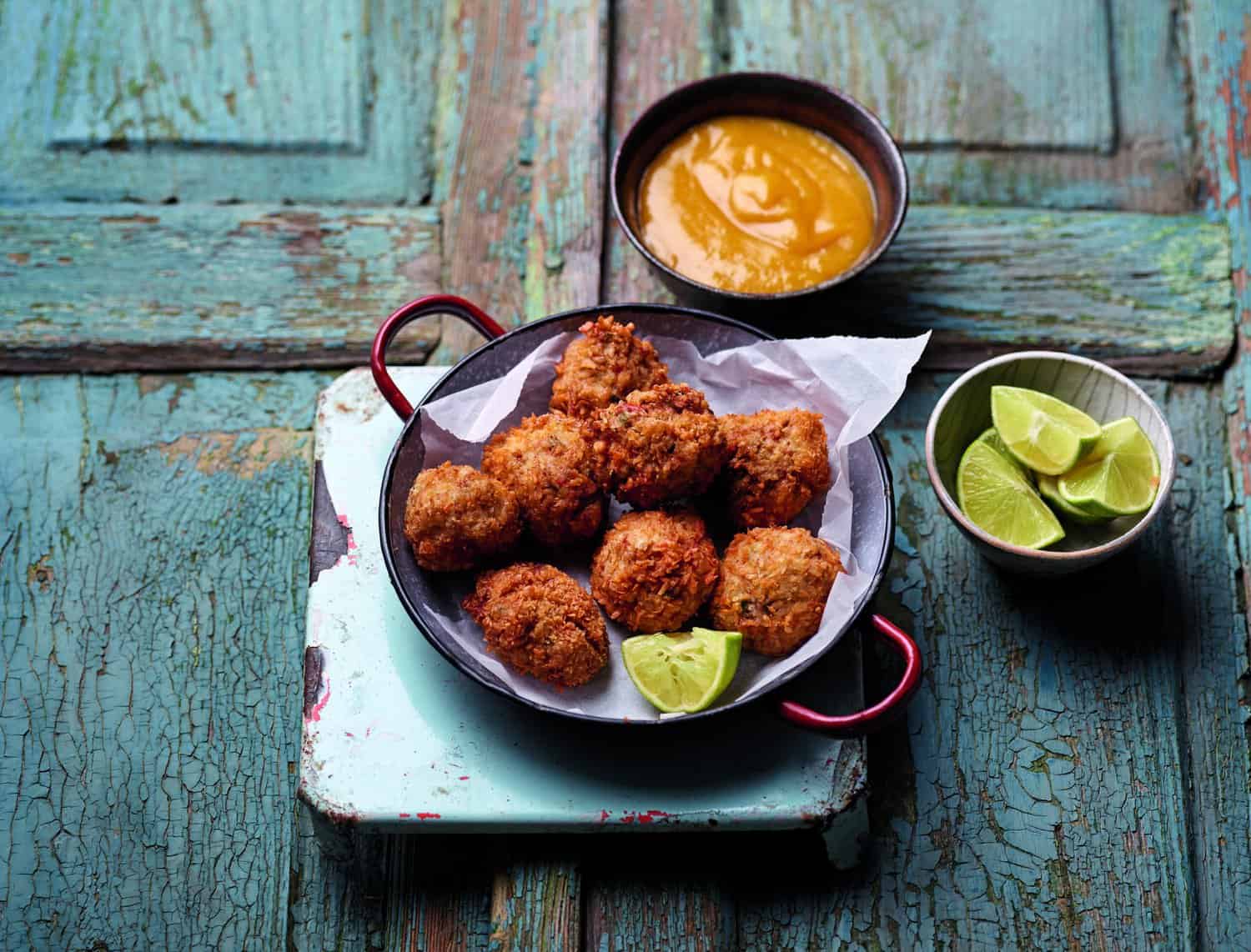 A plate of fritters with a wedge of lime sits on a rustic surface, ready to be the heroes of any gathering. A small bowl of dipping sauce and a bowl with lime wedges are nearby, completing this delightful cooking scene.