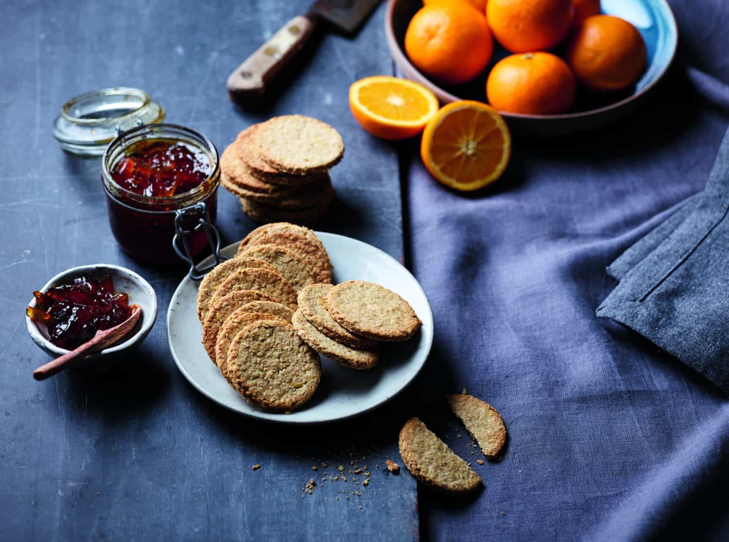 A plate of round crackers, a jar of jam, and a small bowl of sweet spread with a spoon sit like heroes ready for the feast. Nearby, oranges gleam on the dark surface with a gray cloth, embodying culinary delight.