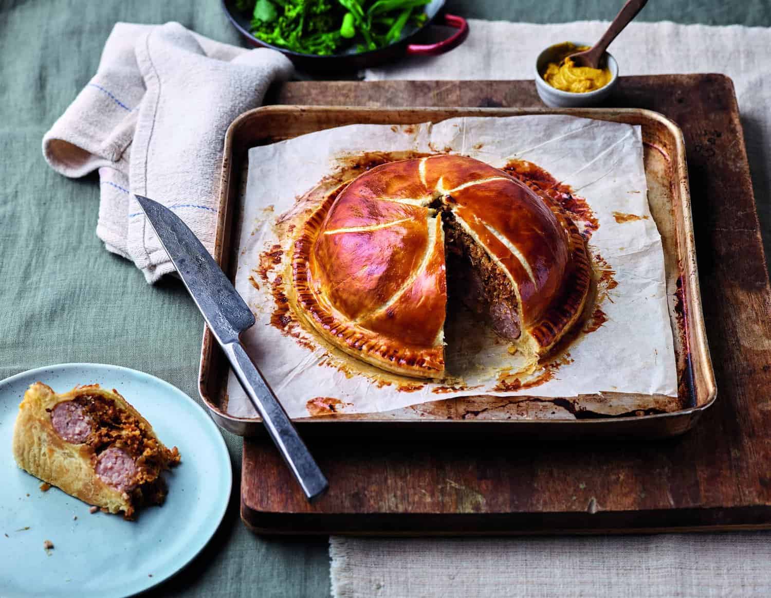 A meat pie on a baking tray with a slice missing reveals its savory filling. A knife rests heroically nearby. The slice is plated beside the tray, ready for tasting. In the background, a pot of mustard and some fresh greenery complete this delightful cooking scene.