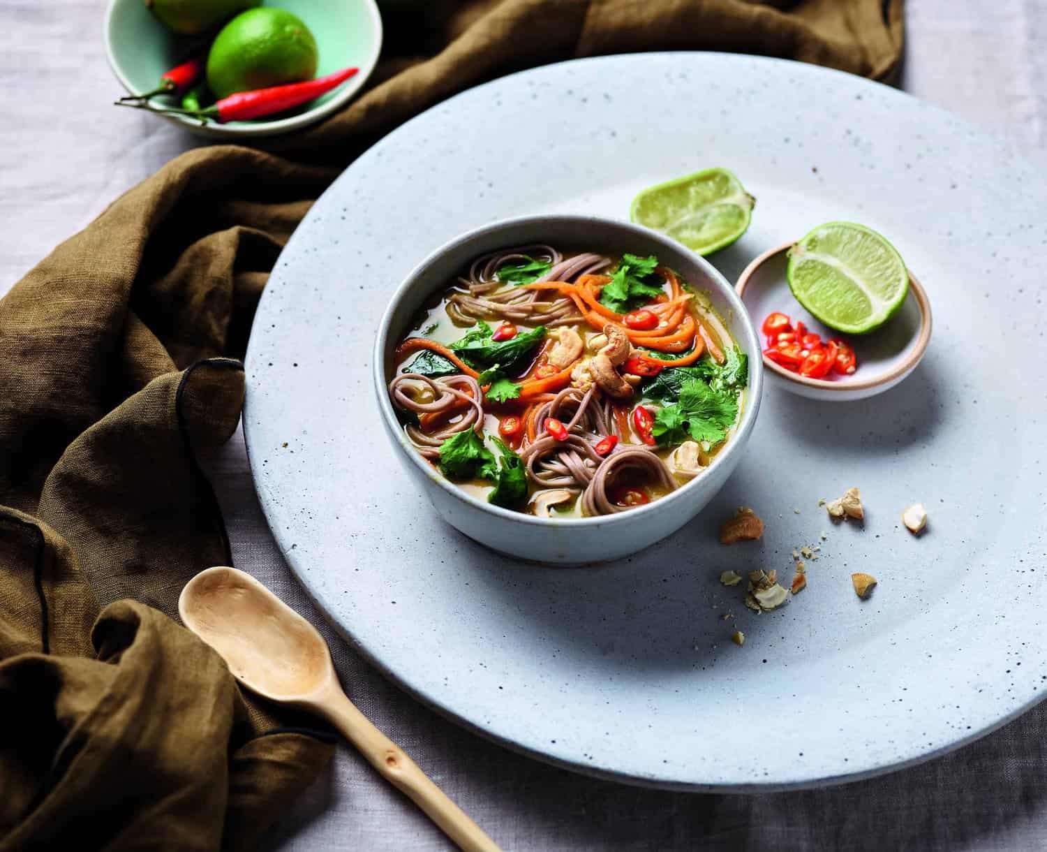 A bowl of colorful noodle soup with vegetables and chicken, garnished with cilantro, is a hero of flavor. Lime wedges and chili slices rest on a small dish beside it. A wooden spoon and brown cloth complete this delightful cooking scene.