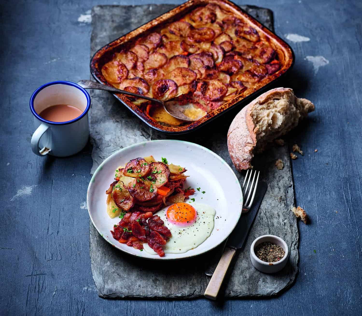 A plate with a fried egg, crispy bacon, and golden potatoes sits heroically beside a baking dish of roasted vegetables. Amidst this culinary delight on the slate surface are a piece of bread, a steaming mug, and a small dish of pepper, turning breakfast into artful cooking.