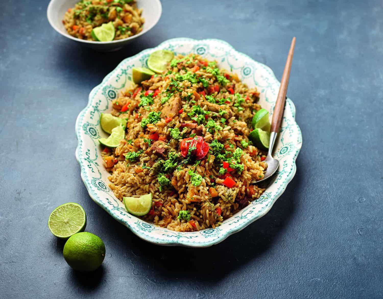 A decorative platter of mixed rice with vegetables and herbs, garnished with lime wedges, showcases the heroes of culinary art on a dark surface. A small bowl with a similar dish and two whole limes complete this cooking masterpiece nearby.