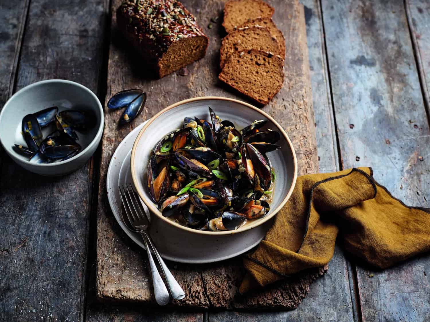 A bowl of cooked mussels with herbs sits heroically on a wooden surface, accompanied by sliced brown bread, a small bowl brimming with more mussels, and a folded brownish cloth.