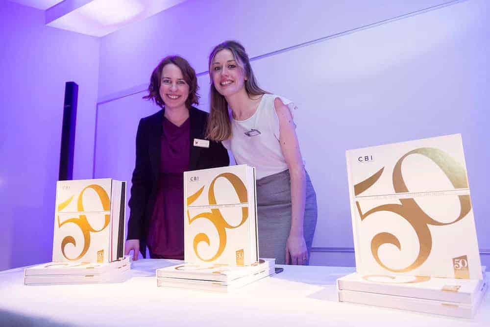 Two women stand behind a table displaying books with "50" on the cover, celebrating 50 years of business innovation under purple lighting.