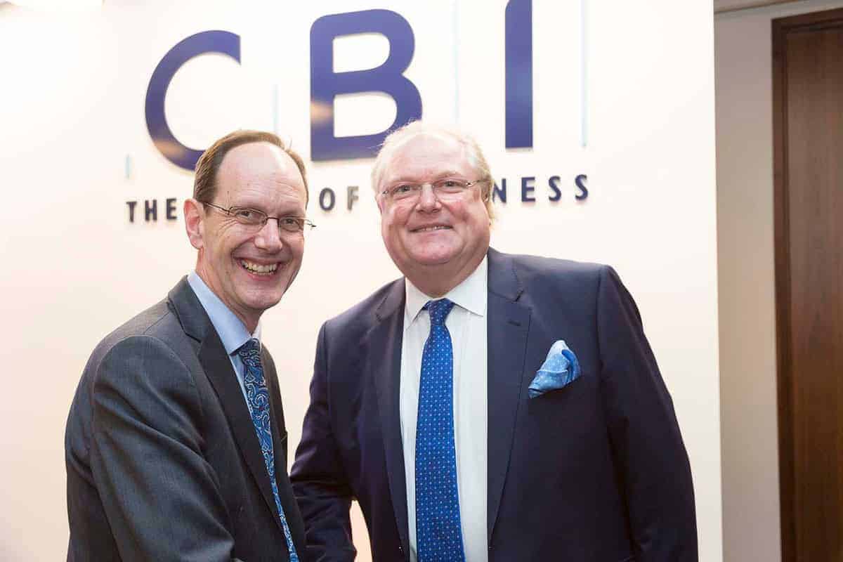 Two men in suits are smiling and shaking hands in front of a CBI sign indoors, celebrating 50 years of business innovation.