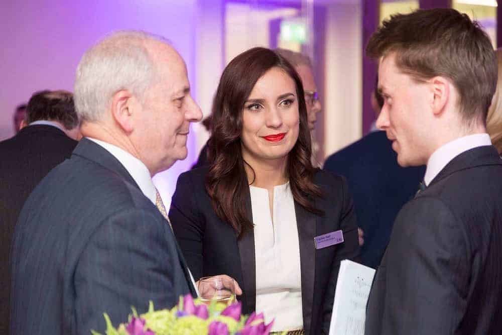 Three people in business attire are engaged in conversation at an indoor event focused on business innovation. A woman in a black jacket is listening intently to two men. A floral arrangement adds elegance to the scene, underscoring the theme of flourishing business growth.