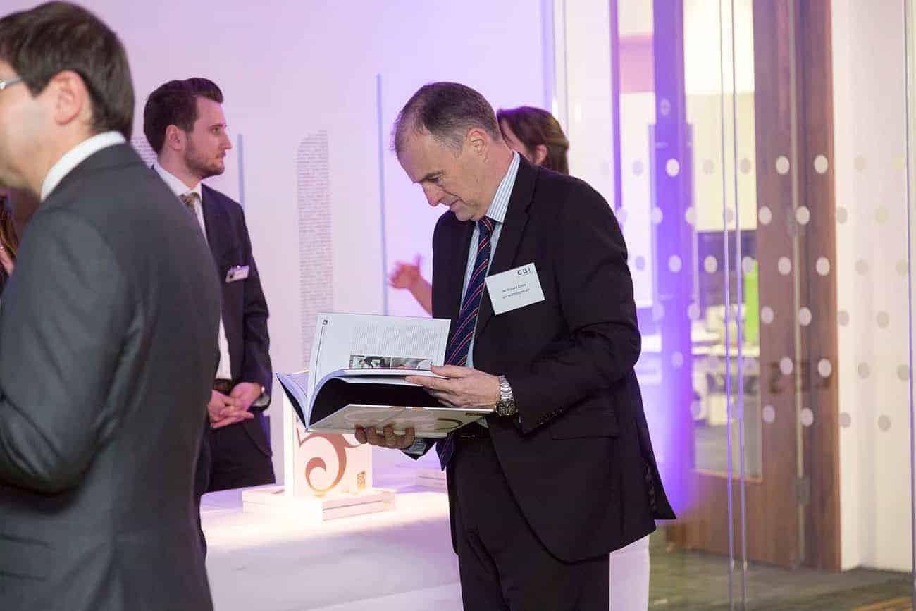 At a formal event celebrating 50 years of business innovation, a man in a suit reads a book, while other attendees engage in discussions visible in the background.