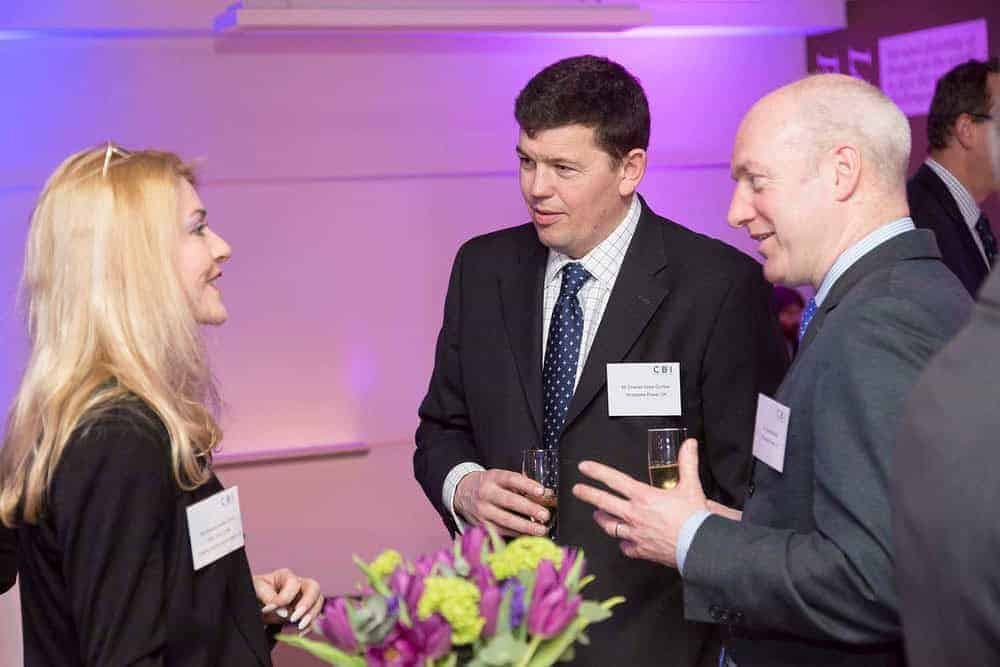 Three people in business attire engage in a conversation while holding drinks, celebrating 50 years of business innovation. In the foreground, a vase of purple flowers adds a touch of elegance to the scene.