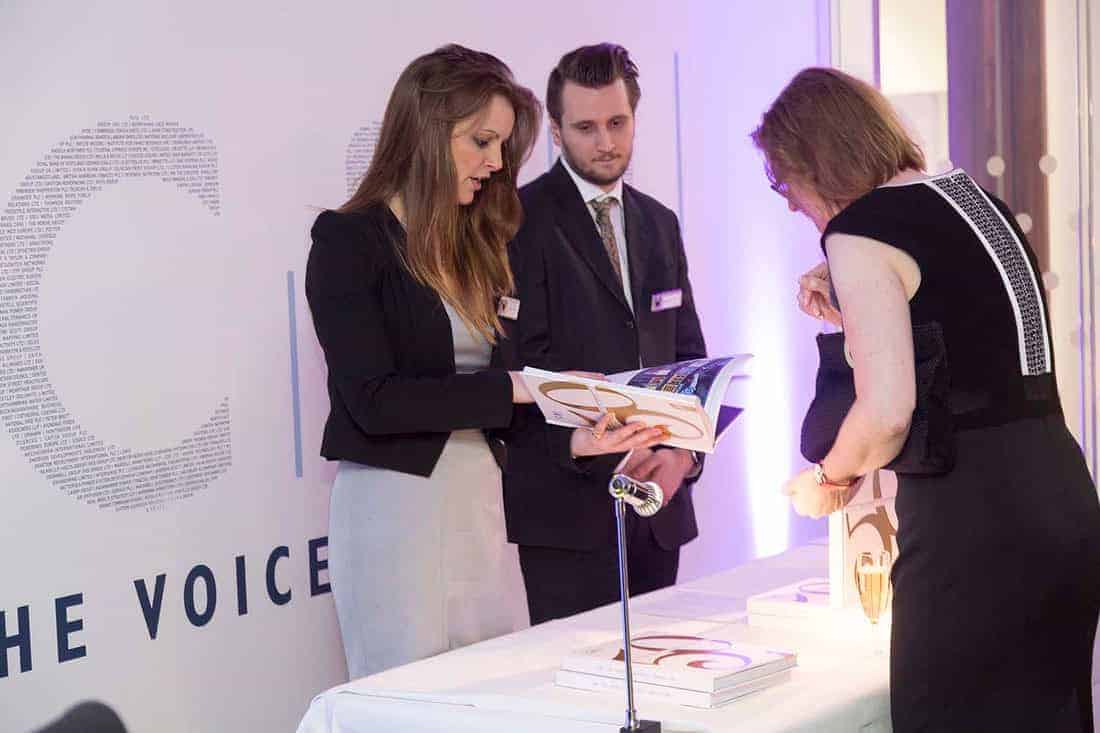 Three people gather at a table with books and a microphone. Two stand while one leans over, viewing a book—a scene reminiscent of celebrating 50 years of business innovation.