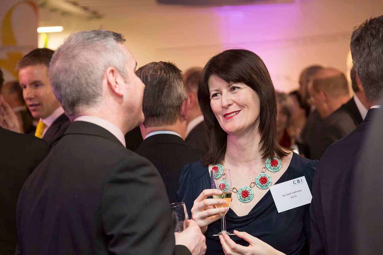 At a formal event celebrating 50 years of business innovation, two people engage in conversation, drinks in hand. A woman wearing a name tag and a decorative necklace listens intently, as other attendees mingle in the background of this grand anniversary gathering.