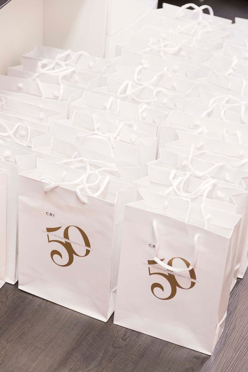 Numerous white gift bags with "50" printed in gold, celebrating 50 years of business innovation, are arranged on the floor, featuring elegant white rope handles.