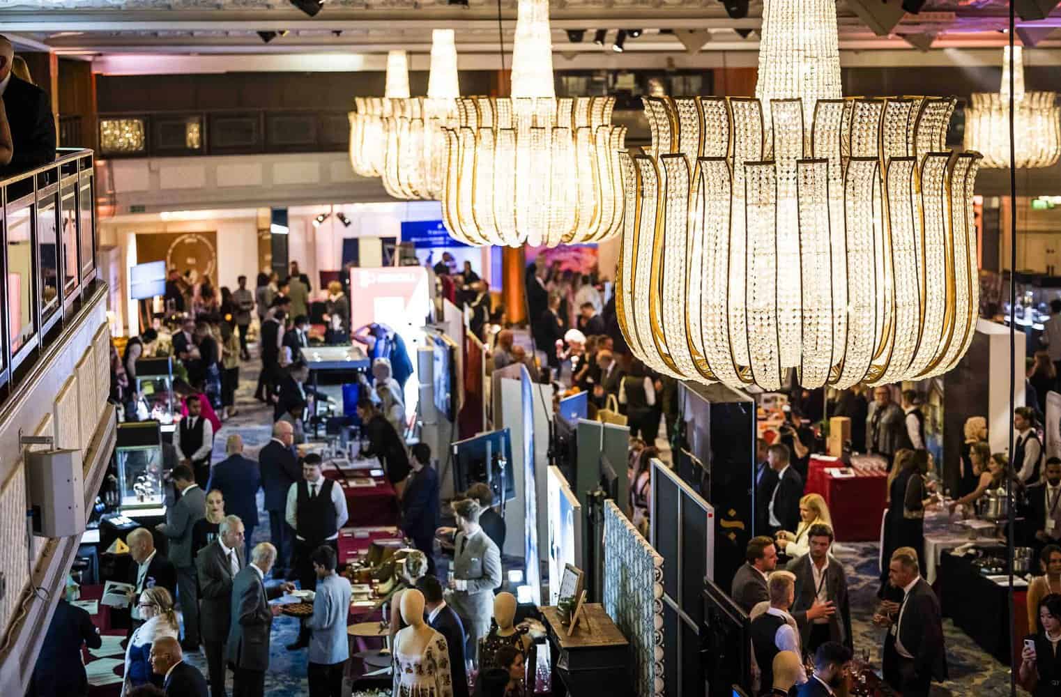 A bustling conference hall with chandeliers, booths, and numerous people striving for perfection while networking and interacting.