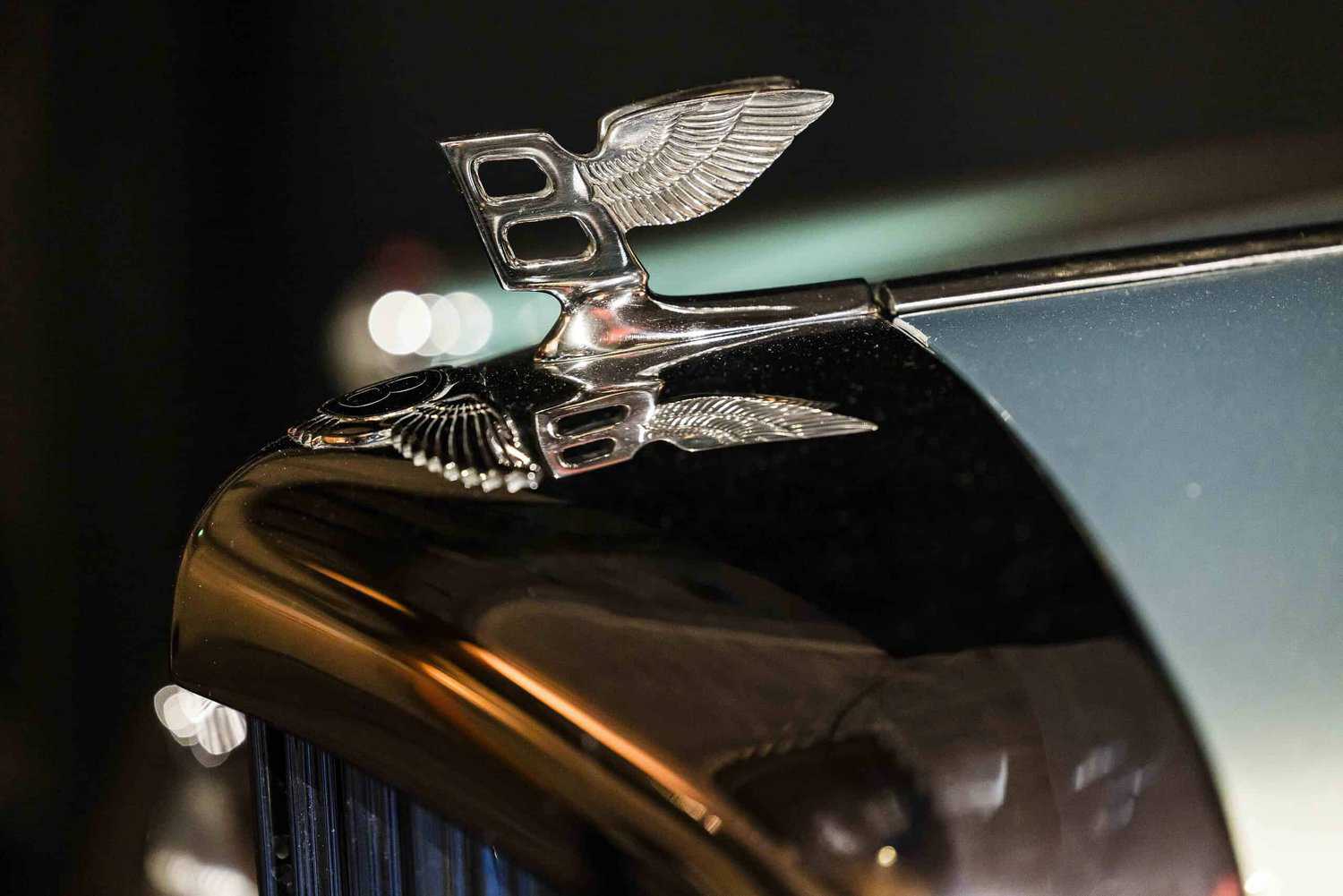 Close-up of a car emblem featuring a silver winged "B" on a metallic surface, embodying the brand's ethos to strive for perfection. The front grille is partially visible, highlighting exceptional craftsmanship.