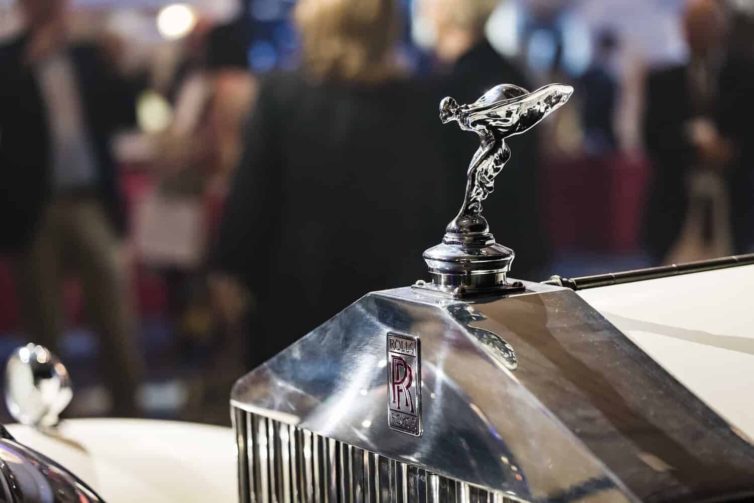 Close-up of a Rolls-Royce hood ornament, the Spirit of Ecstasy, striving for perfection as it graces the car's grille with blurred people in the background.