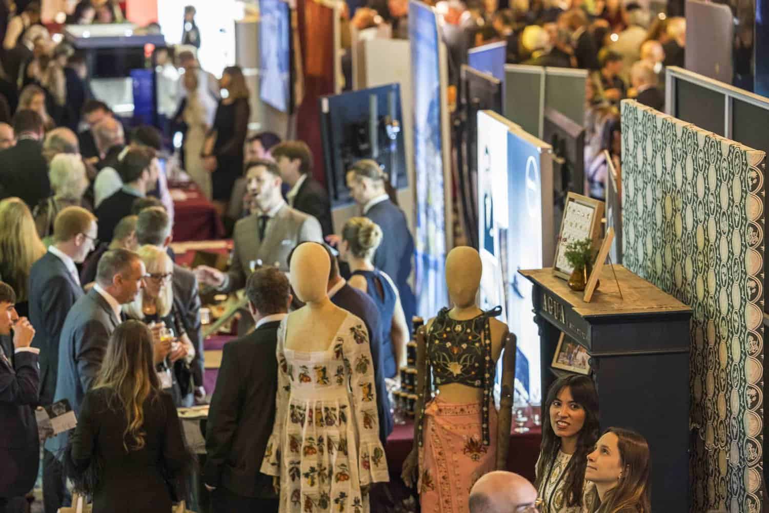A crowded indoor event buzzes with conversation as mannequins artfully display garments, embodying a strive for perfection. Booths and displays line the room, while attendees in formal and casual attire mingle seamlessly.