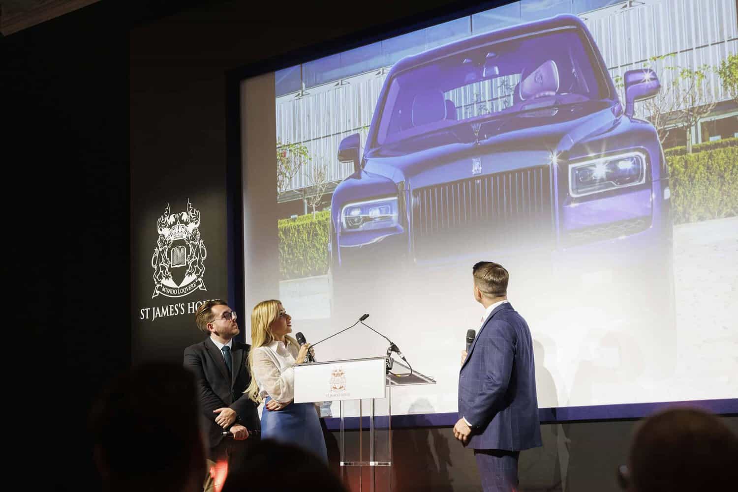 Three people stand on stage, admiring a projected image of a purple car on the screen at St James's House event, embodying the spirit to strive for perfection.