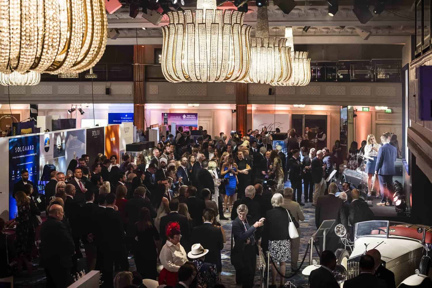 A large crowd in formal attire mingles at an indoor event beneath ornate chandeliers, embodying a strive for perfection. Display booths and a classic car are visible in the background, adding an elegant touch.