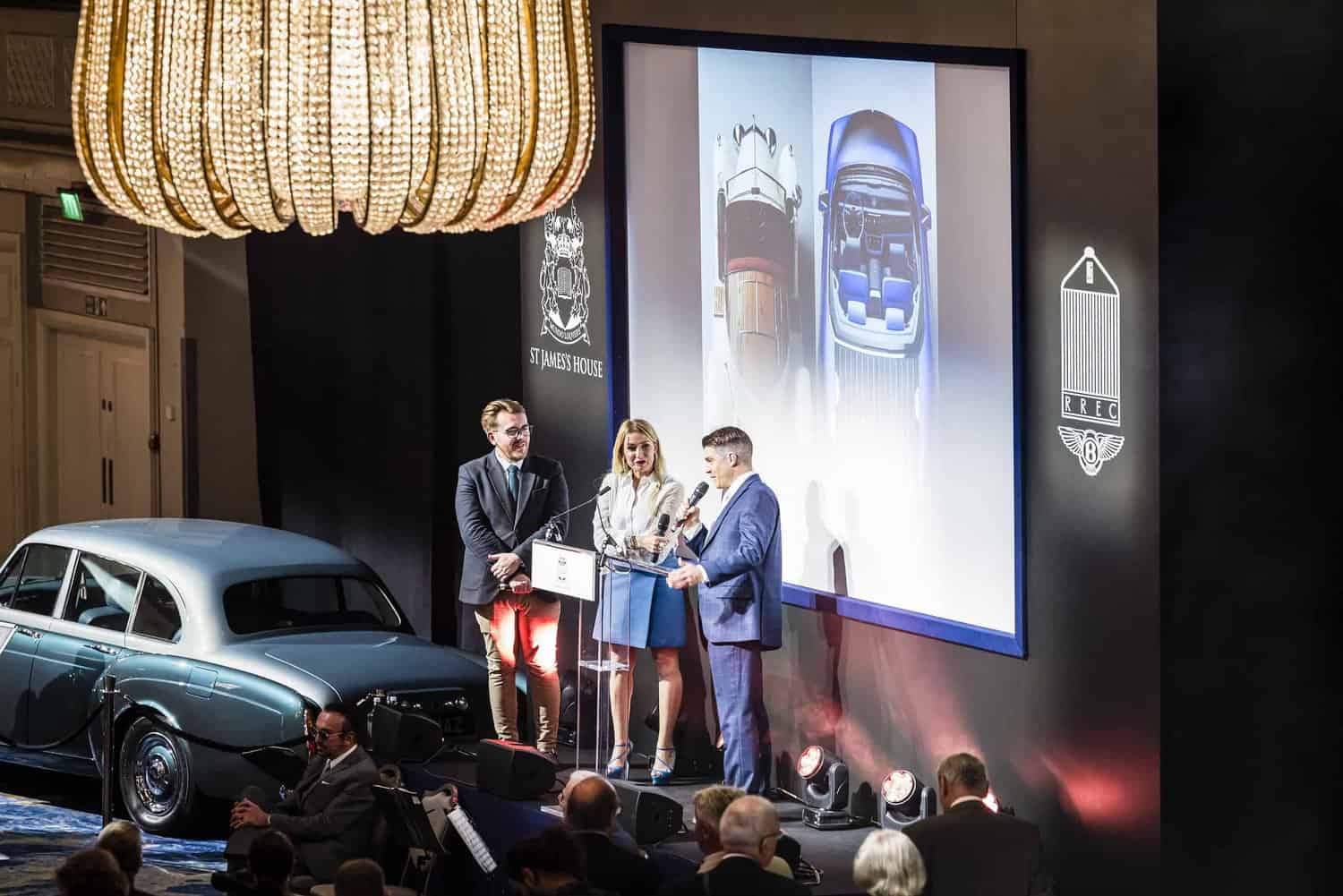 At an event where people stand on stage presenting a vintage car image, with an audience attentively seated, the classic car displayed to the side embodies a timeless pursuit: Strive for Perfection.