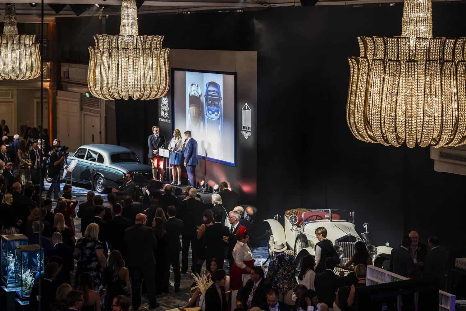 A crowded classic car auction event, graced by two large chandeliers, sees enthusiasts striving for perfection. The scene buzzes with people mingling and admired cars on display. On stage, speakers share insights on a presentation screen, adding to the evening's allure.