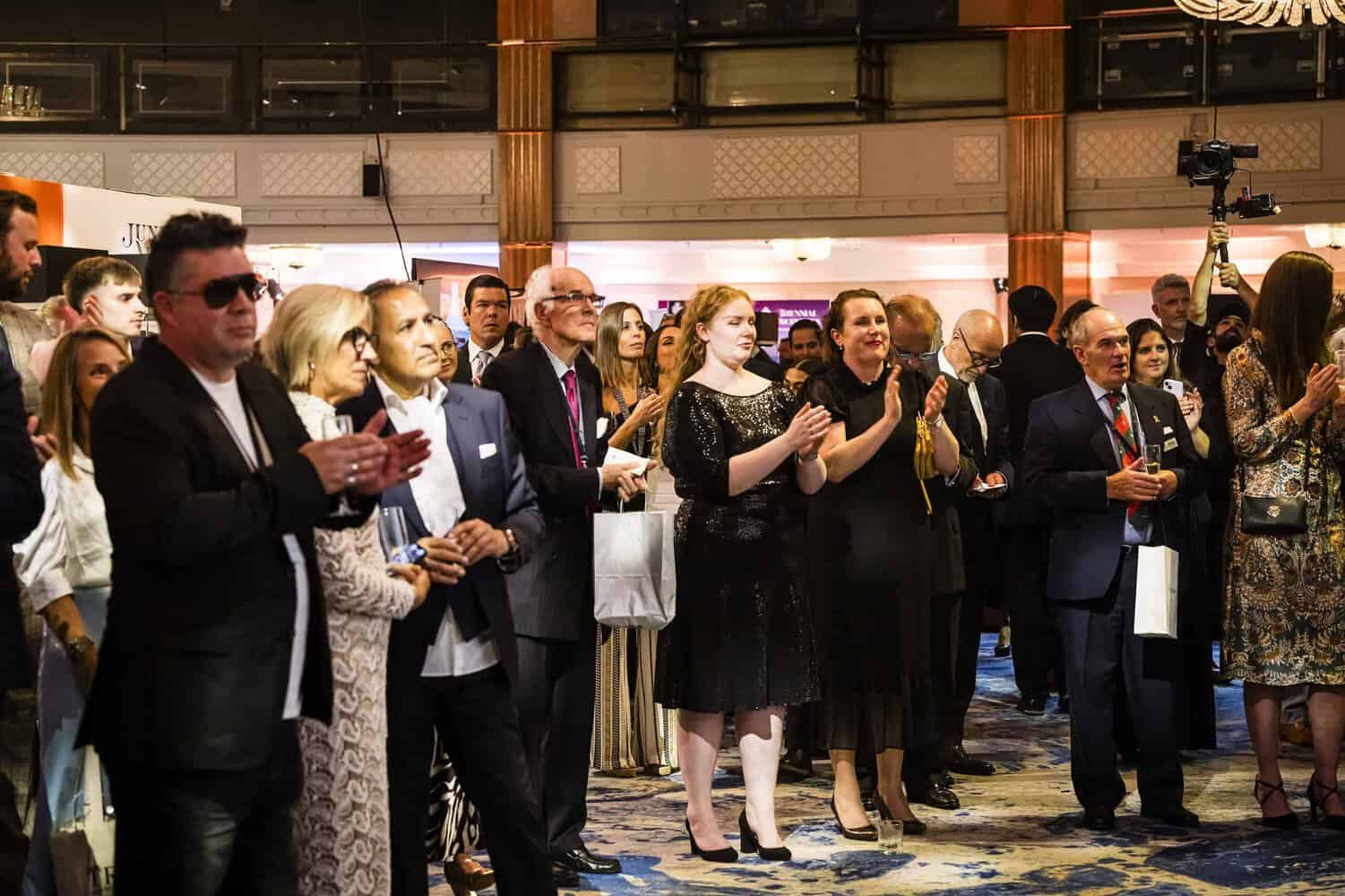 In a large decorated room, a group of people dressed in formal attire stand and applaud, embodying the perfection they strive for.