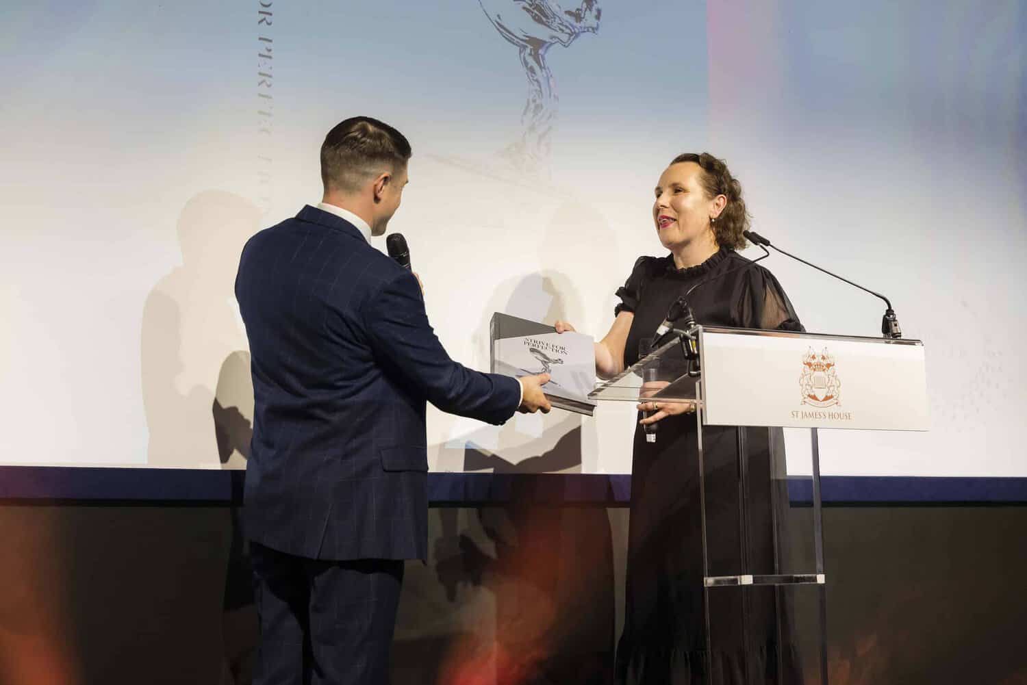 A woman in a black dress, embodying perfection, receives an award on stage from a man in a suit holding a microphone.