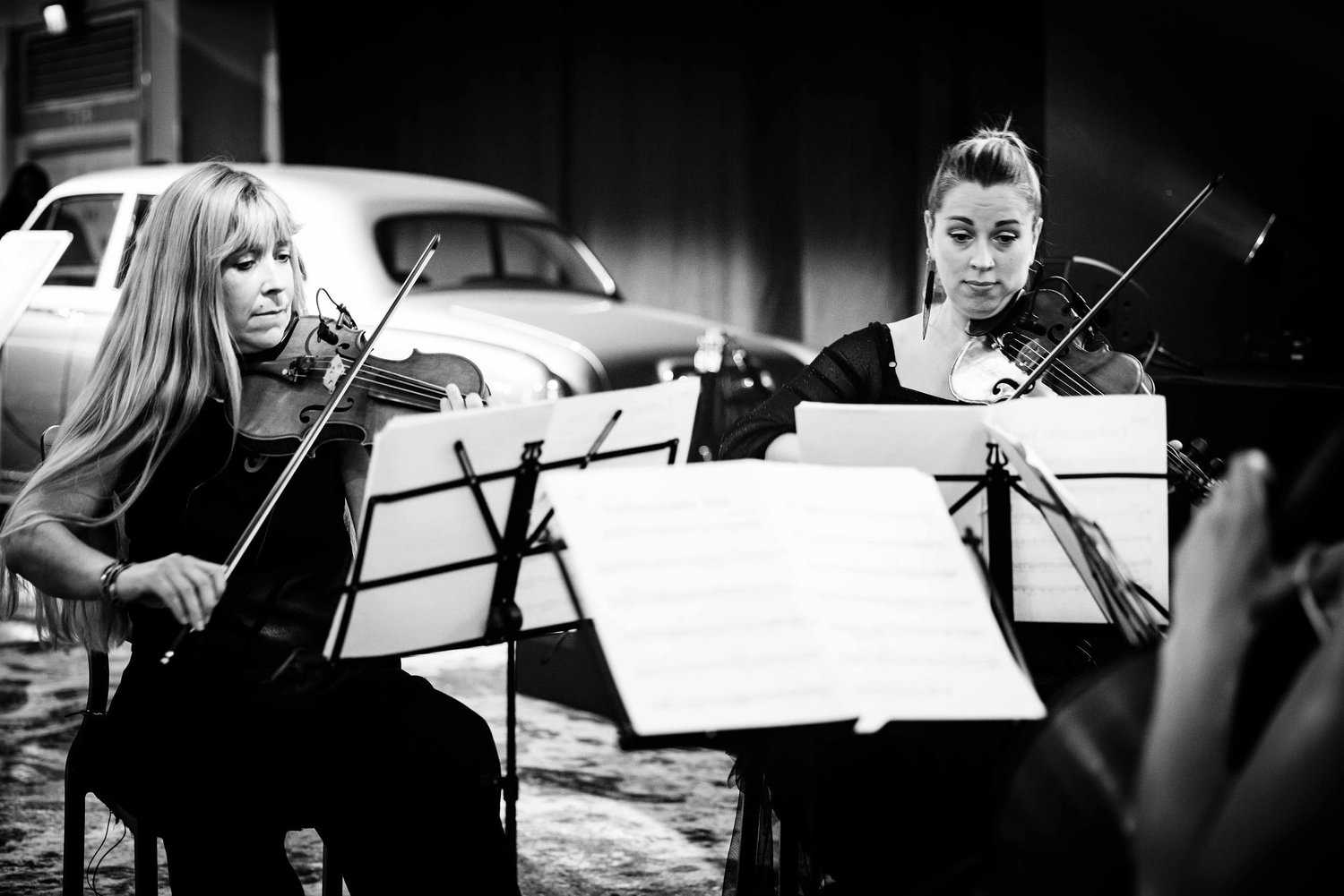 Two women play violins in a string ensemble, seated with music stands and sheet music, exuding perfection. A classic car is visible in the background of this black and white photo.