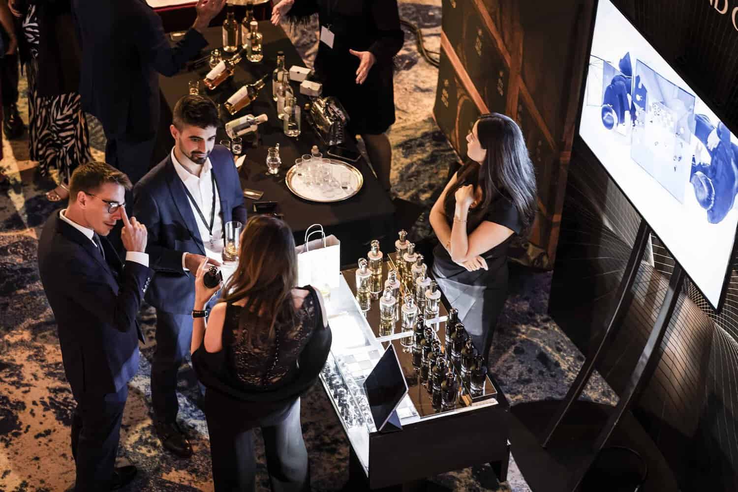 People gathered around a booth with bottles and a screen at an event, engaged in conversation and examining products, all while striving for perfection in their quest for the best.
