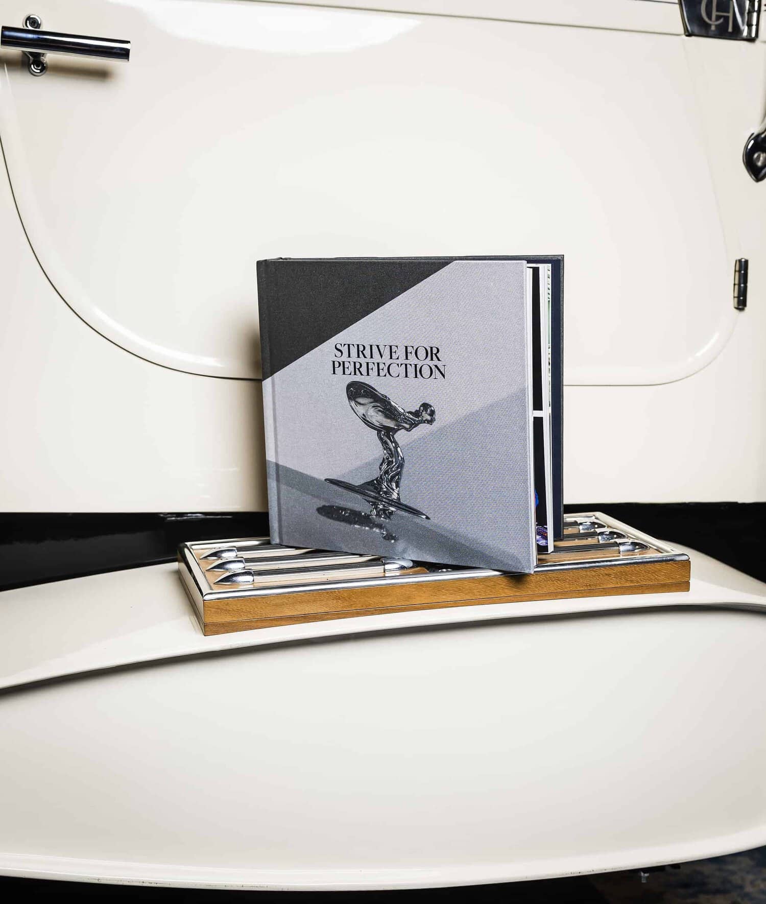 A book titled "Strive for Perfection" delicately rests on a tray with cutlery, all elegantly arranged atop a vintage car door.