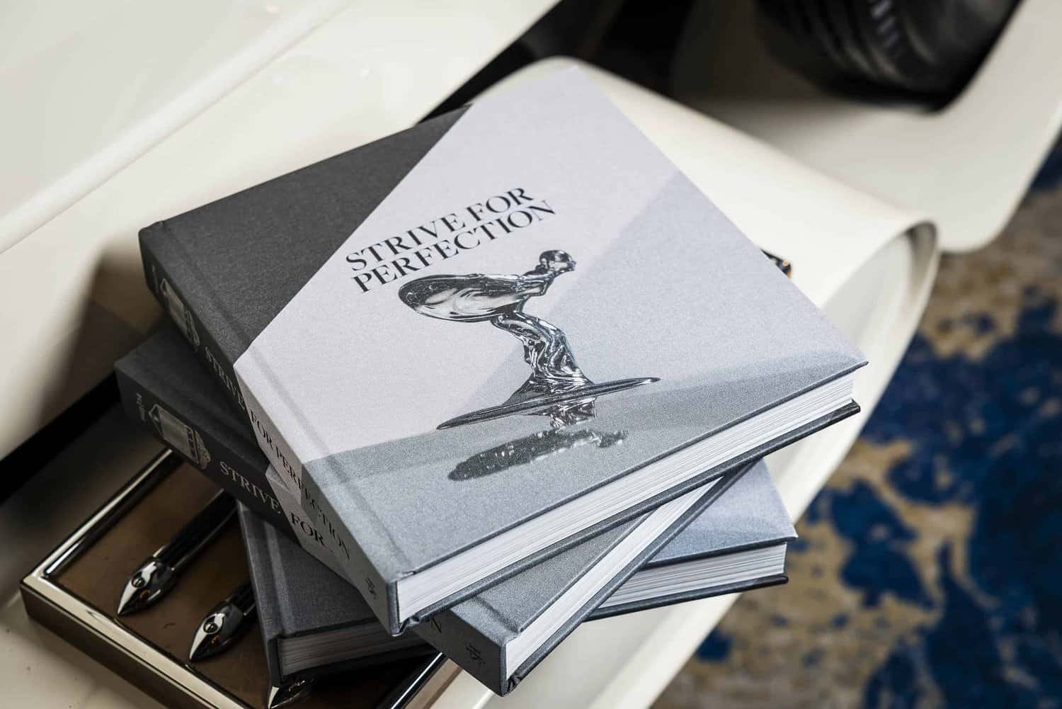 A stack of books titled "Strive for Perfection" with a polished silver figurine on the cover sits elegantly on a pristine white surface.