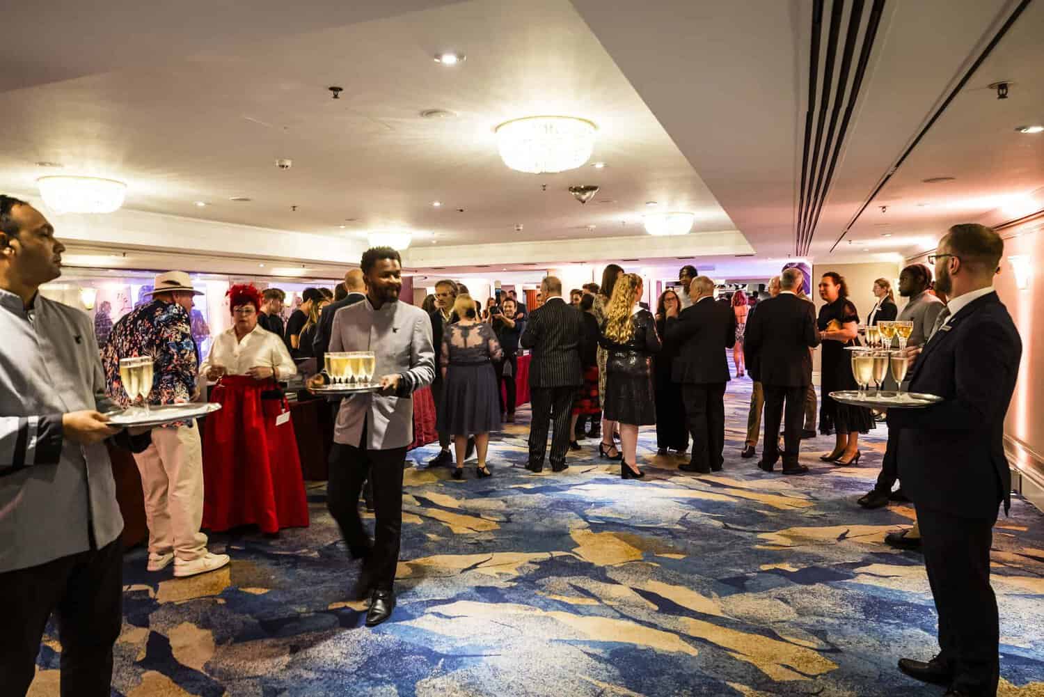 A group of people socializes at a formal event in a well-lit room, their conversations and laughter mingling with the clinking of glasses as servers elegantly glide by, striving for perfection with trays of drinks in hand.