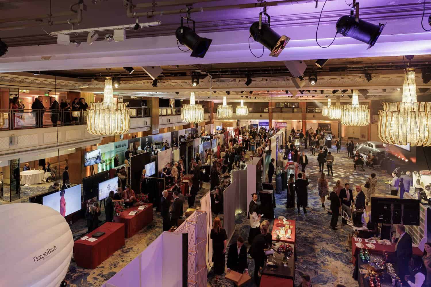 A bustling indoor exhibition with various booths, where attendees strive for connections and inspiration, walking around under the glow of large chandeliers overhead.