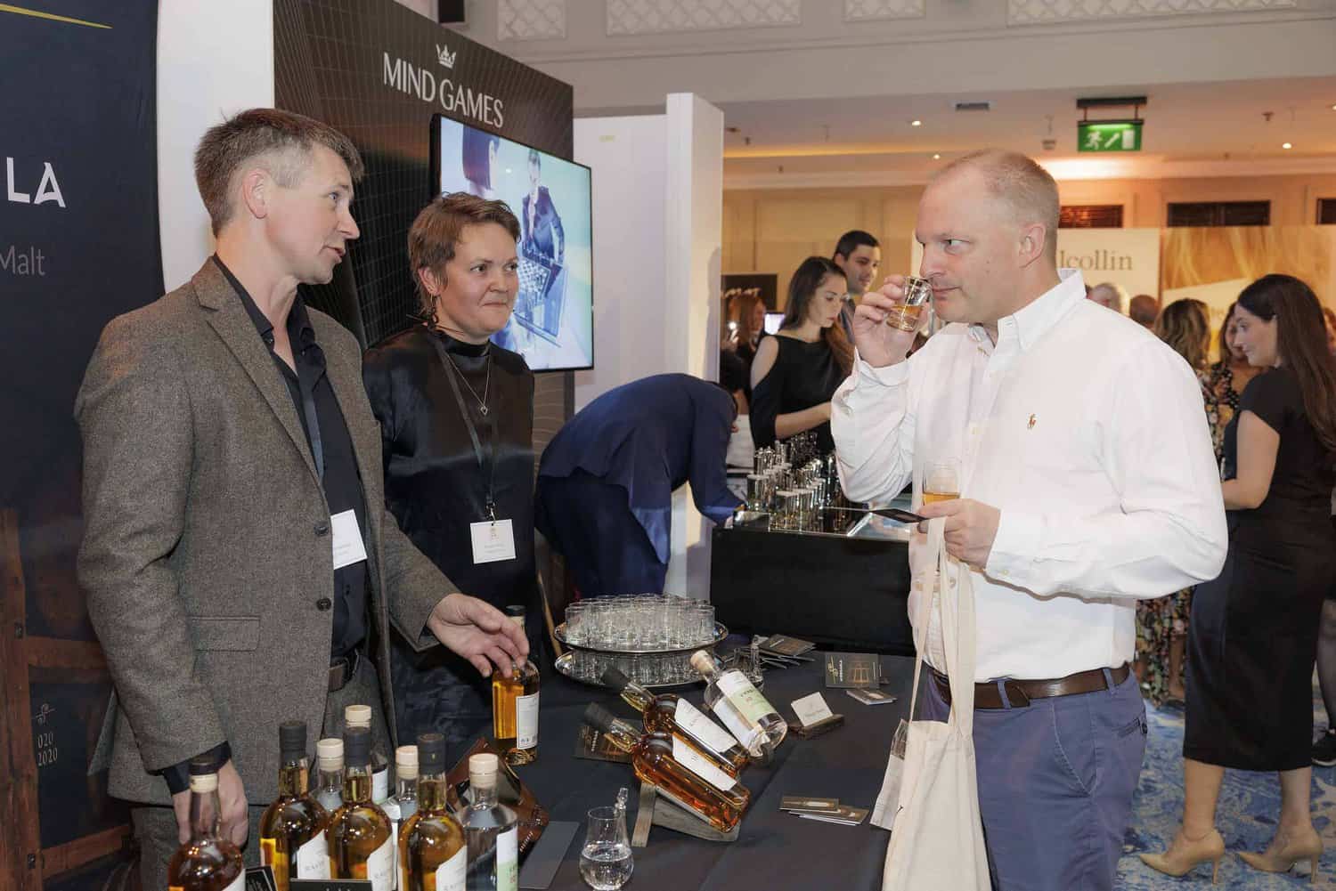 A man samples a drink at a booth with two representatives during an event, savoring the perfection of flavors among the array of liquor bottles displayed on the table.