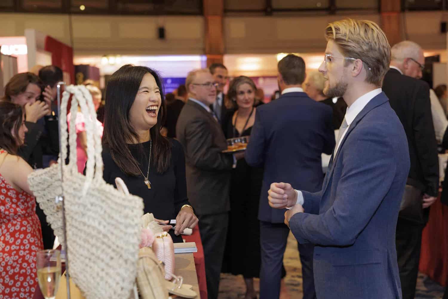 Amidst the bustling crowd of the indoor event, two people are engrossed in conversation. The woman is smiling warmly, her demeanor reflecting a strive for perfection. Nearby, a beautifully crafted decorative item graces the table, adding elegance to their engaging exchange.