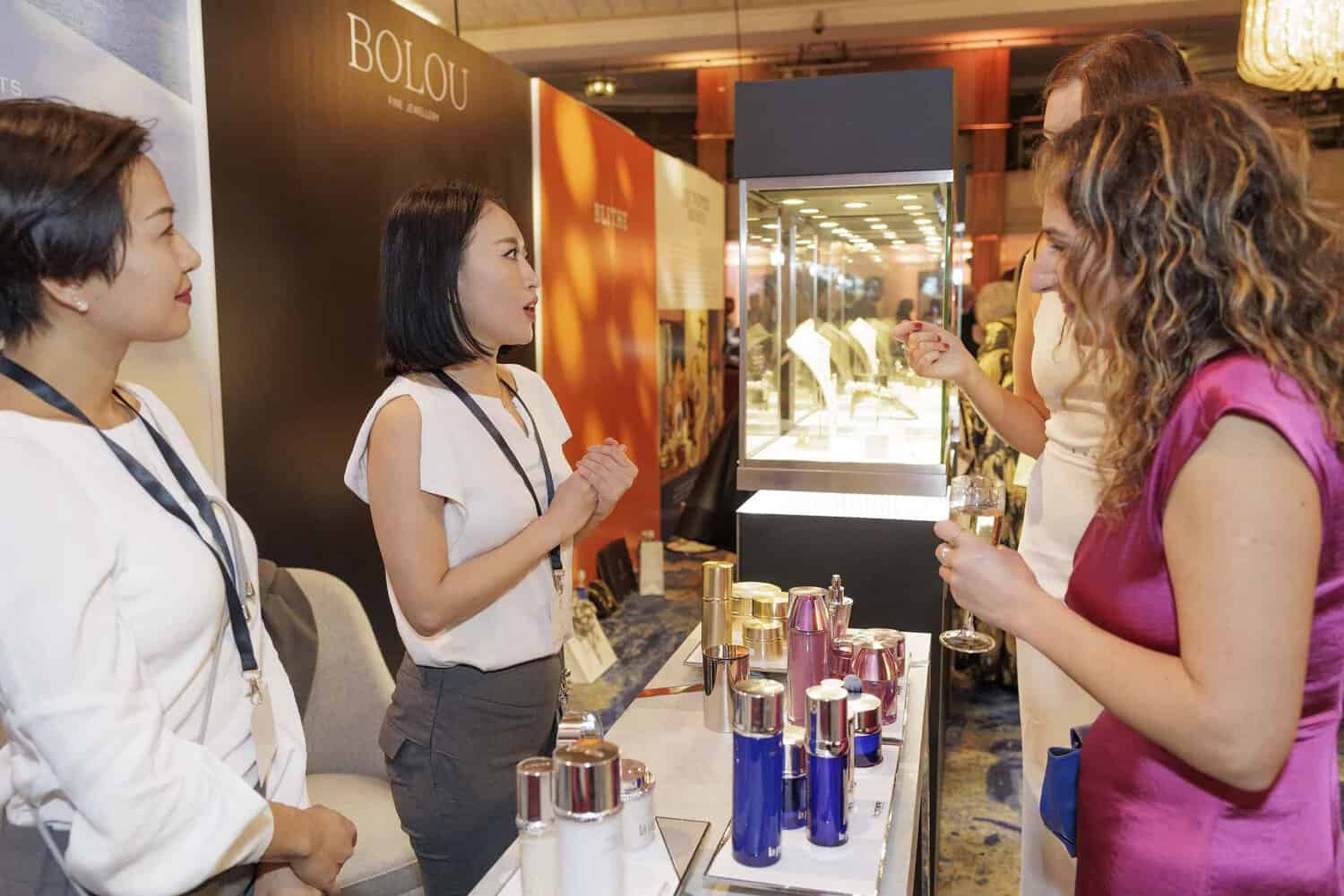 Two women stand at a display table adorned with cosmetic products, striving for perfection in every detail as they engage in conversation with two other women holding glasses.