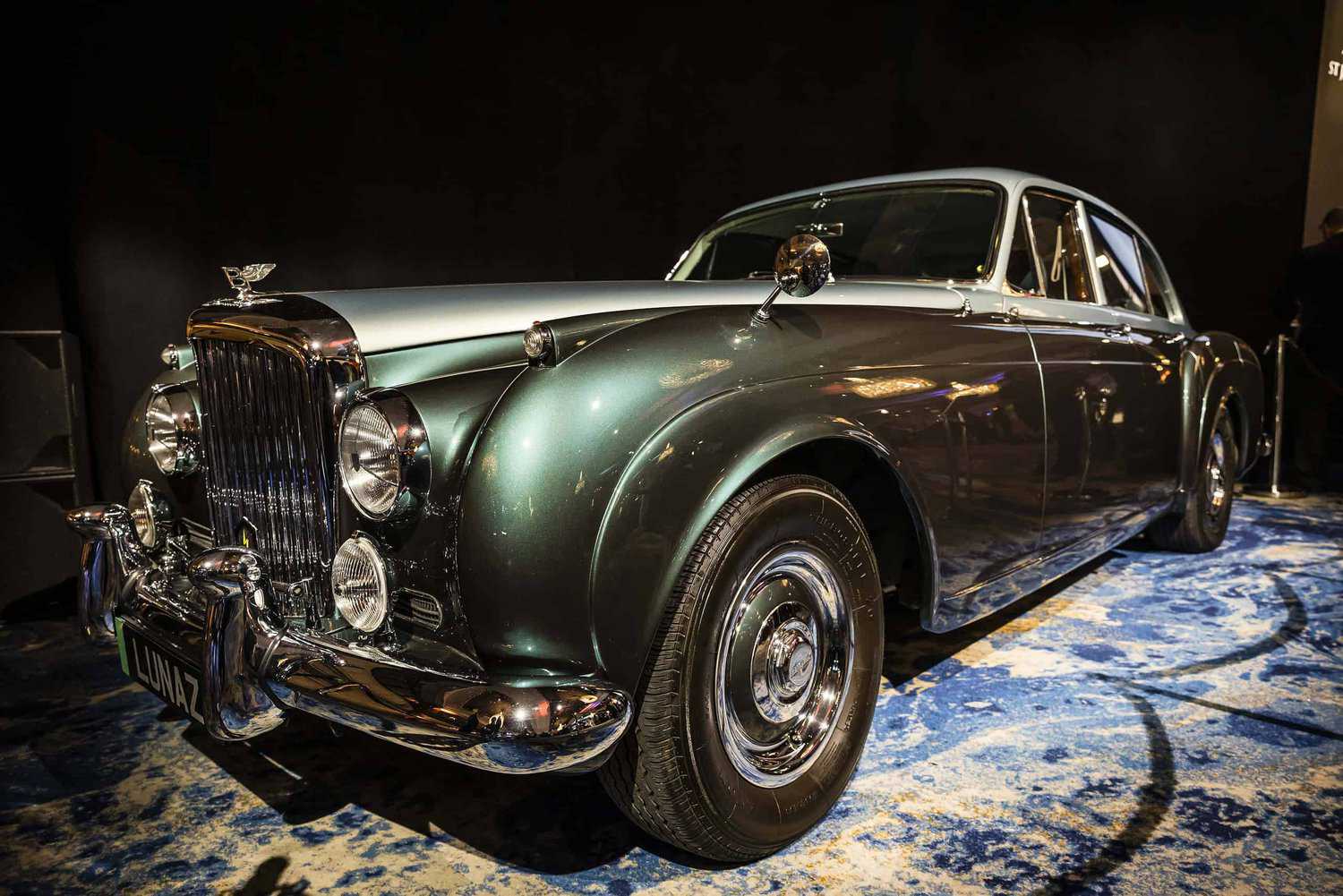 In a dimly lit showroom, a vintage green and silver luxury car gleams on a blue-patterned carpet—a testament to those who strive for perfection.
