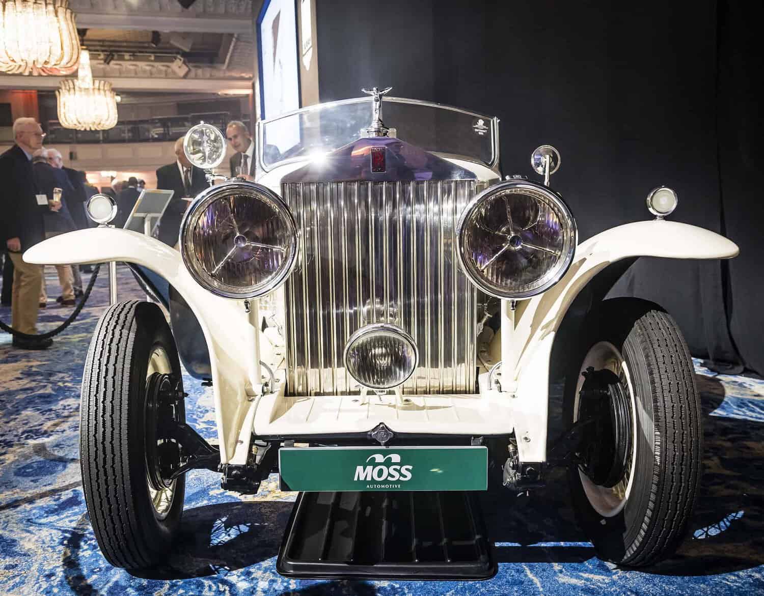Front view of a vintage white car displayed indoors with a "Moss" sign at the front, where perfection meets style, featuring prominent headlights, a classic grille, and a shiny finish.